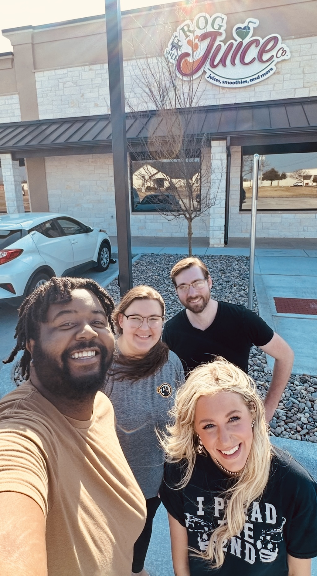 Four people smiling outside a building with a sign that reads 'Frog Juice,' parked cars in the background.