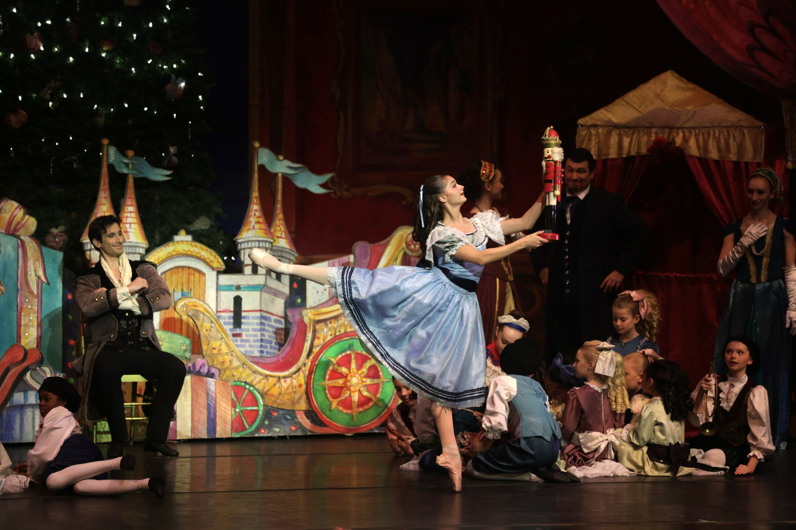 Group of children and adults performing a ballet on stage with colorful set pieces and a Christmas tree in the background.