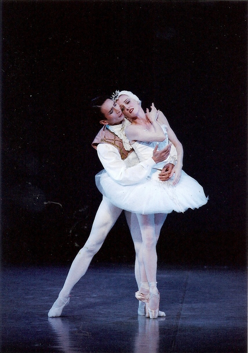 Two ballet dancers perform on stage, one supporting the other in a pose, with a dark background.