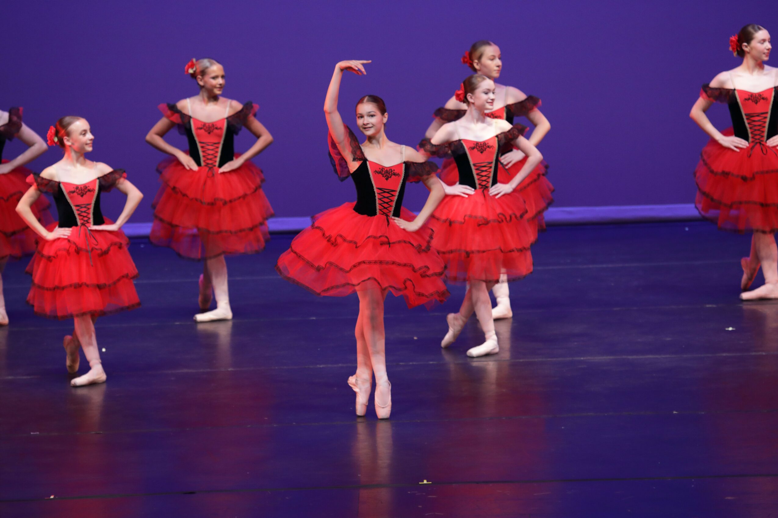 Group of young ballet dancers in red and black costumes performing on stage with purple background.