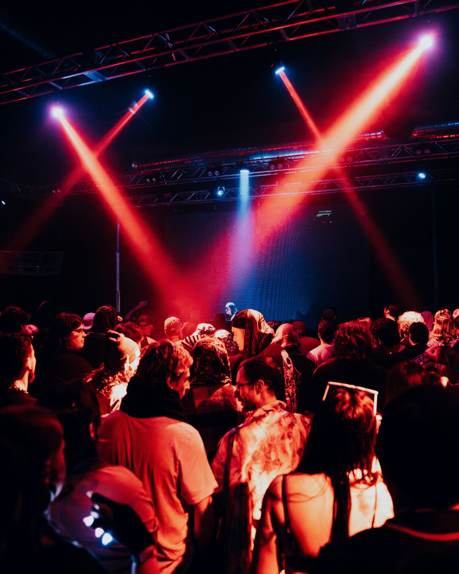 Crowd watching a concert with colorful stage lights and beams shining down, audience facing the stage.