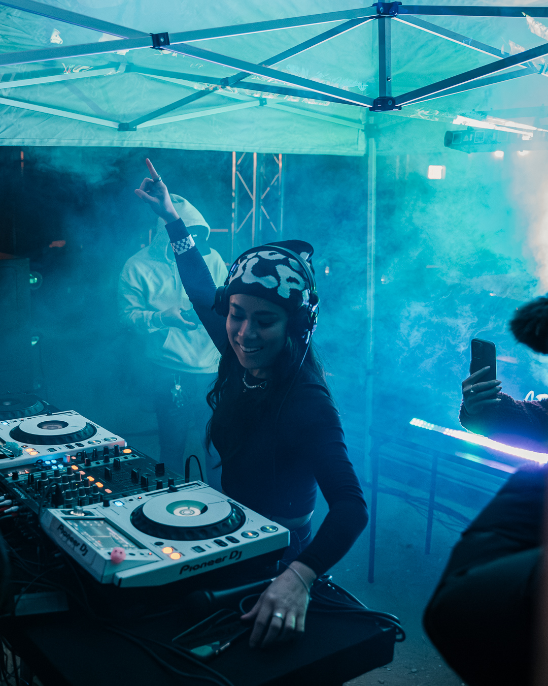 Woman DJ wearing a patterned headscarf, smiling, raising her hand, with DJ equipment and blue lighting in background.
