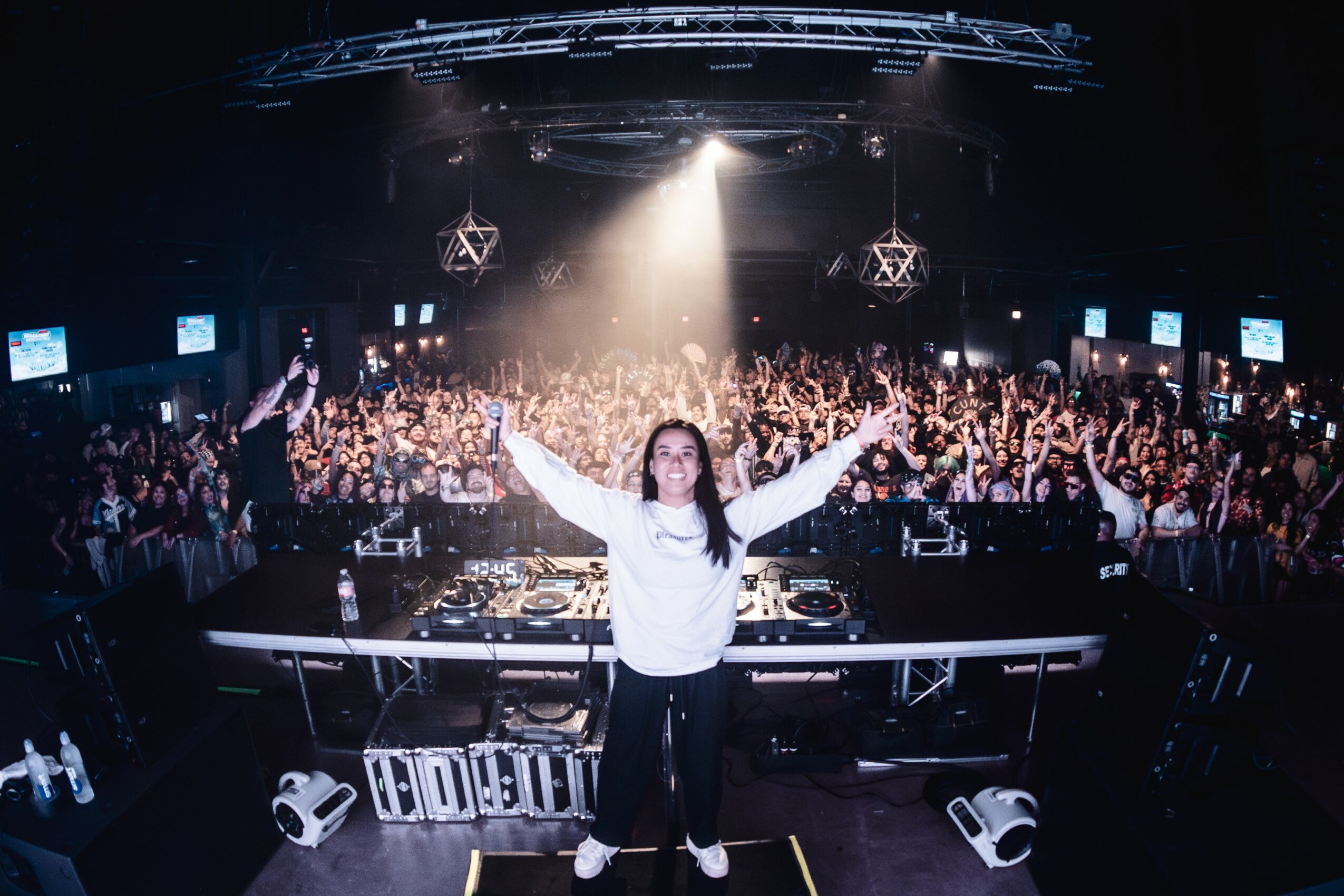 Person standing with arms raised in front of a large crowd at a concert or event, stage and screens visible behind.