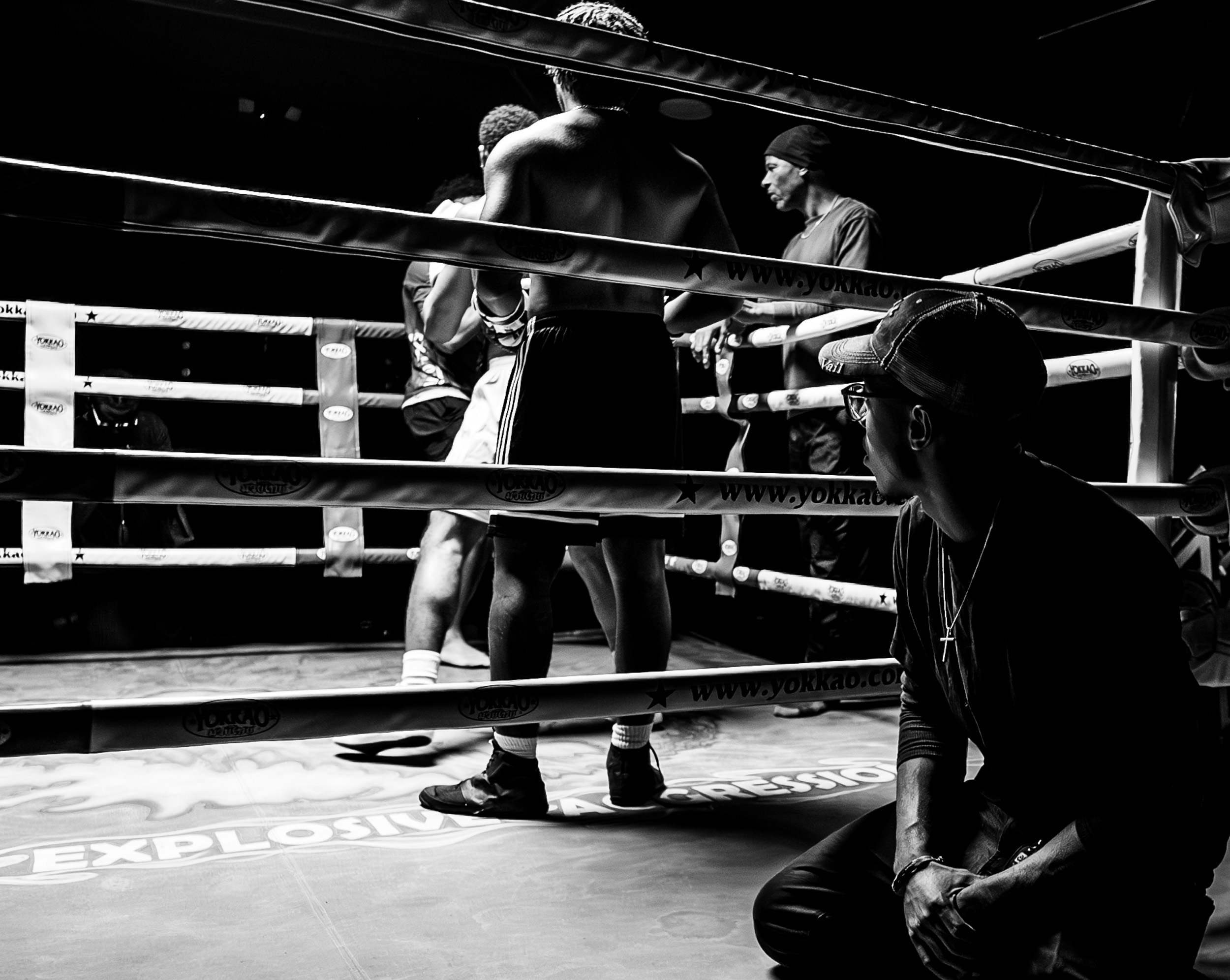 Boxers and a coach inside a boxing ring, with one boxer resting and the coach sitting nearby, in black and white.