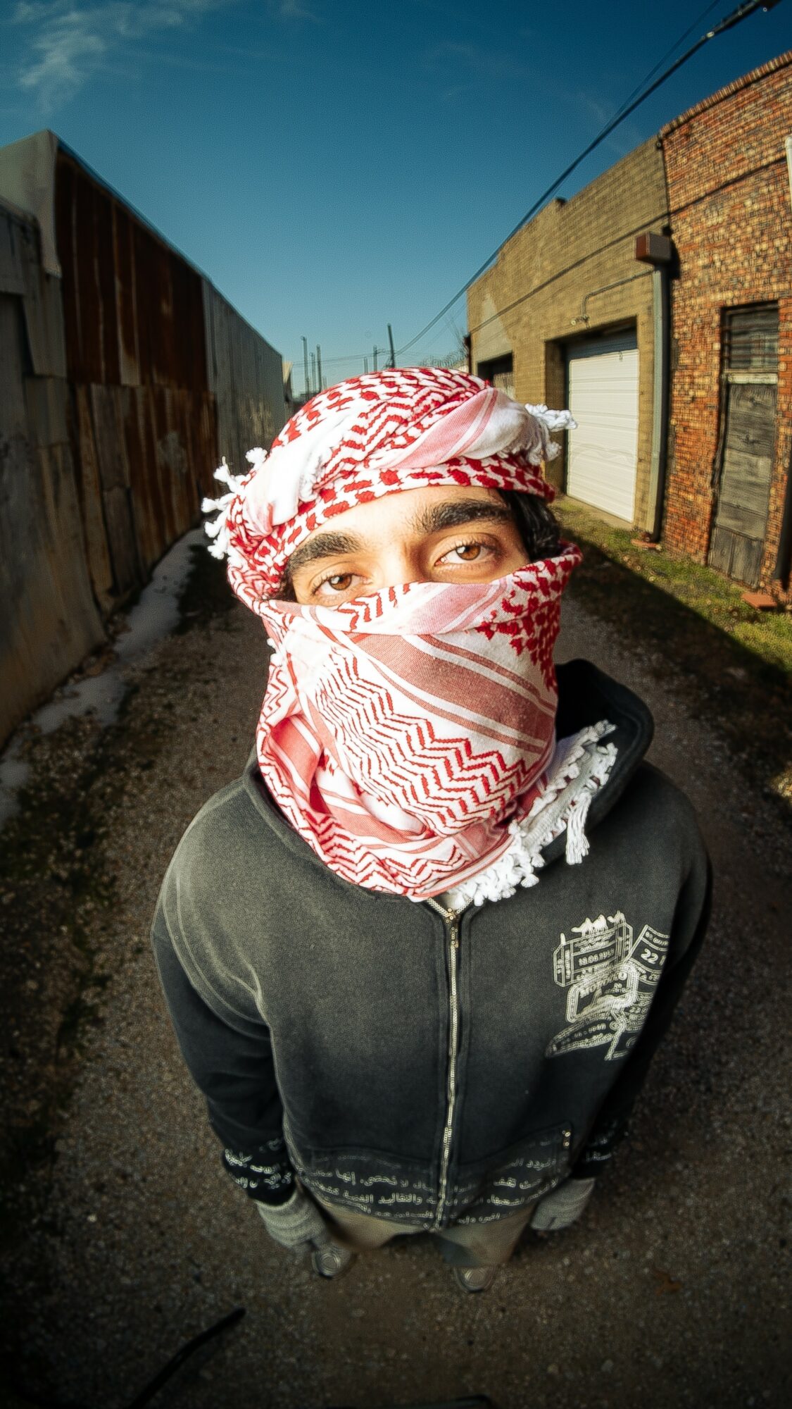 Person wearing a red and white patterned head covering and face mask, standing outdoors in an alleyway with brick and metal walls.
