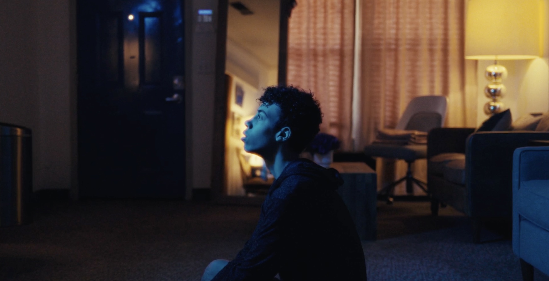 Person with face painted blue sitting in a dimly lit room, looking to the side, with furniture and a lamp in background.