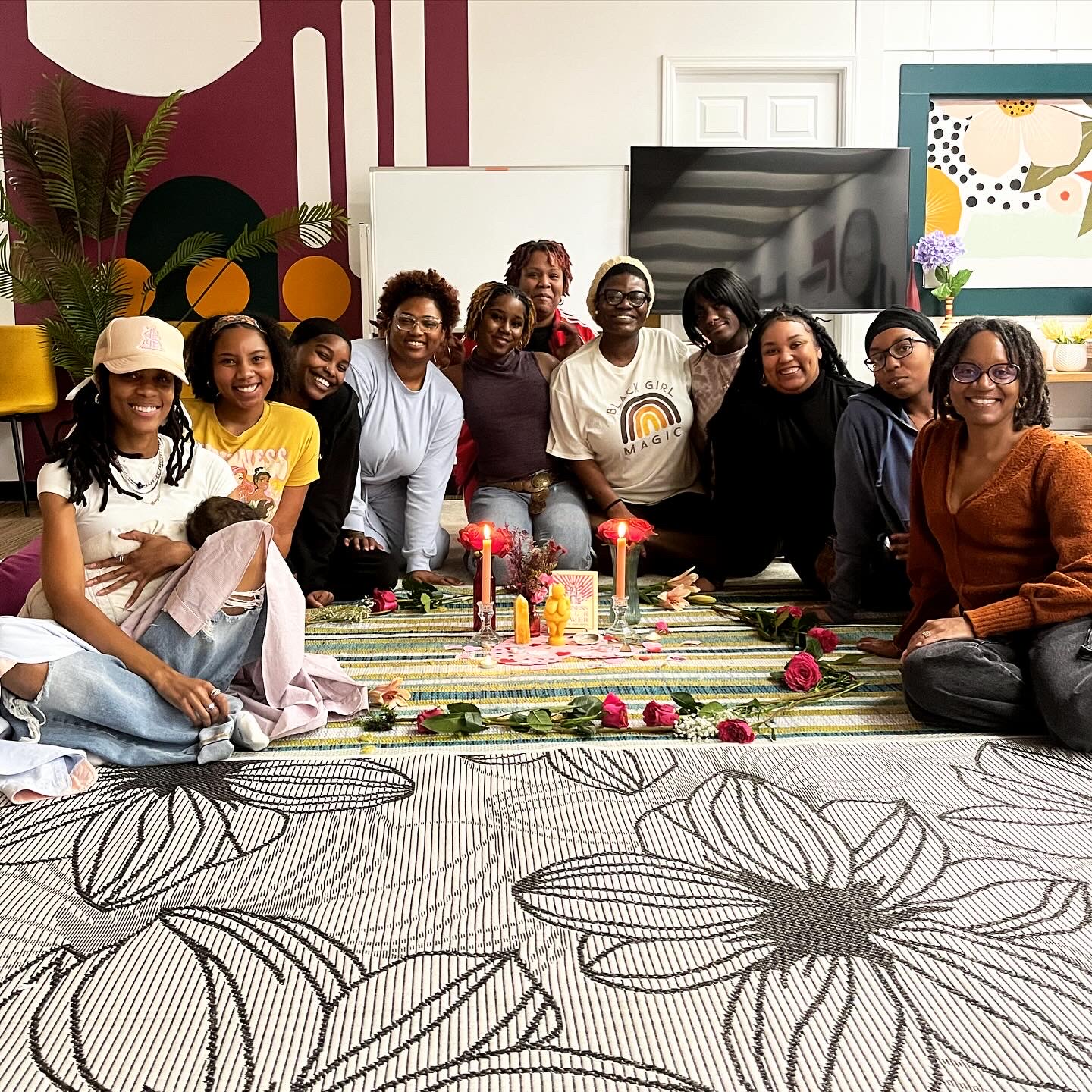 Group of people sitting on a colorful rug with candles and flowers, smiling indoors with artwork and plants in background.