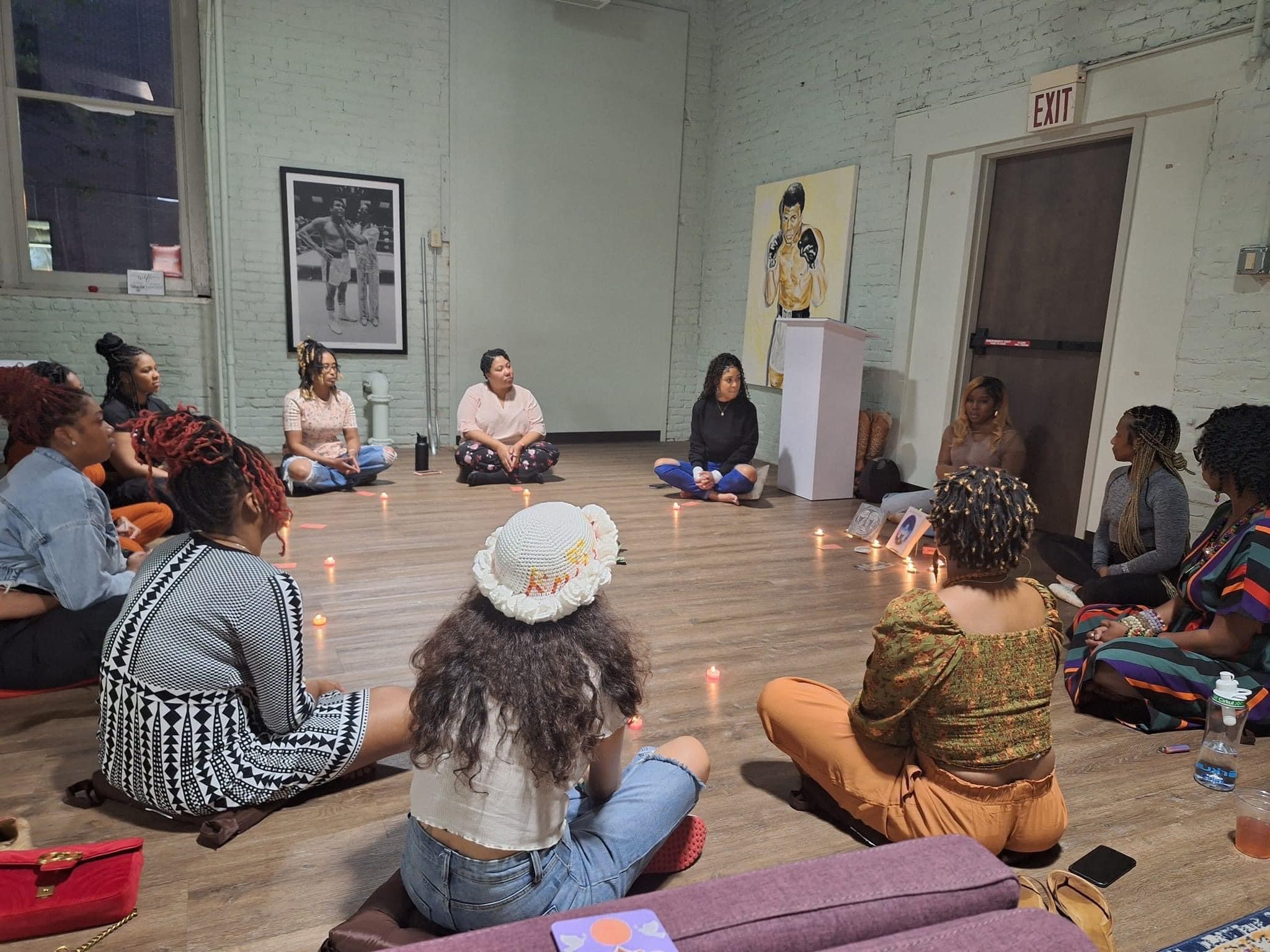 Group of people sitting in a circle on the floor in a room with artwork on the walls, candles, and a water bottle.