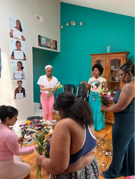 Group of women and a girl gather around flowers in a room with teal and light green walls, some holding flowers.