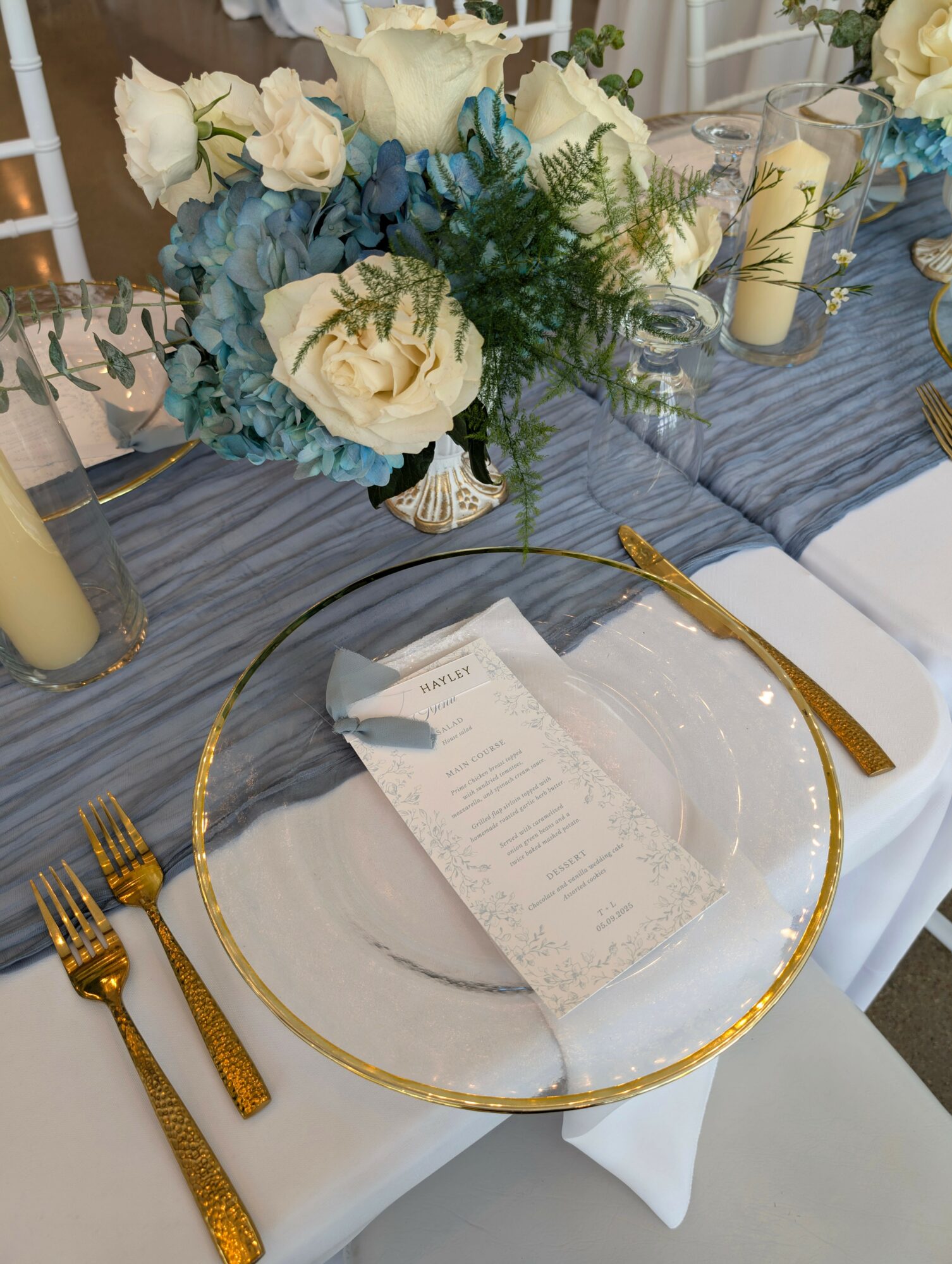 Elegant table setting with a floral centerpiece, gold utensils, and a menu on a white napkin, with a blue tablecloth.