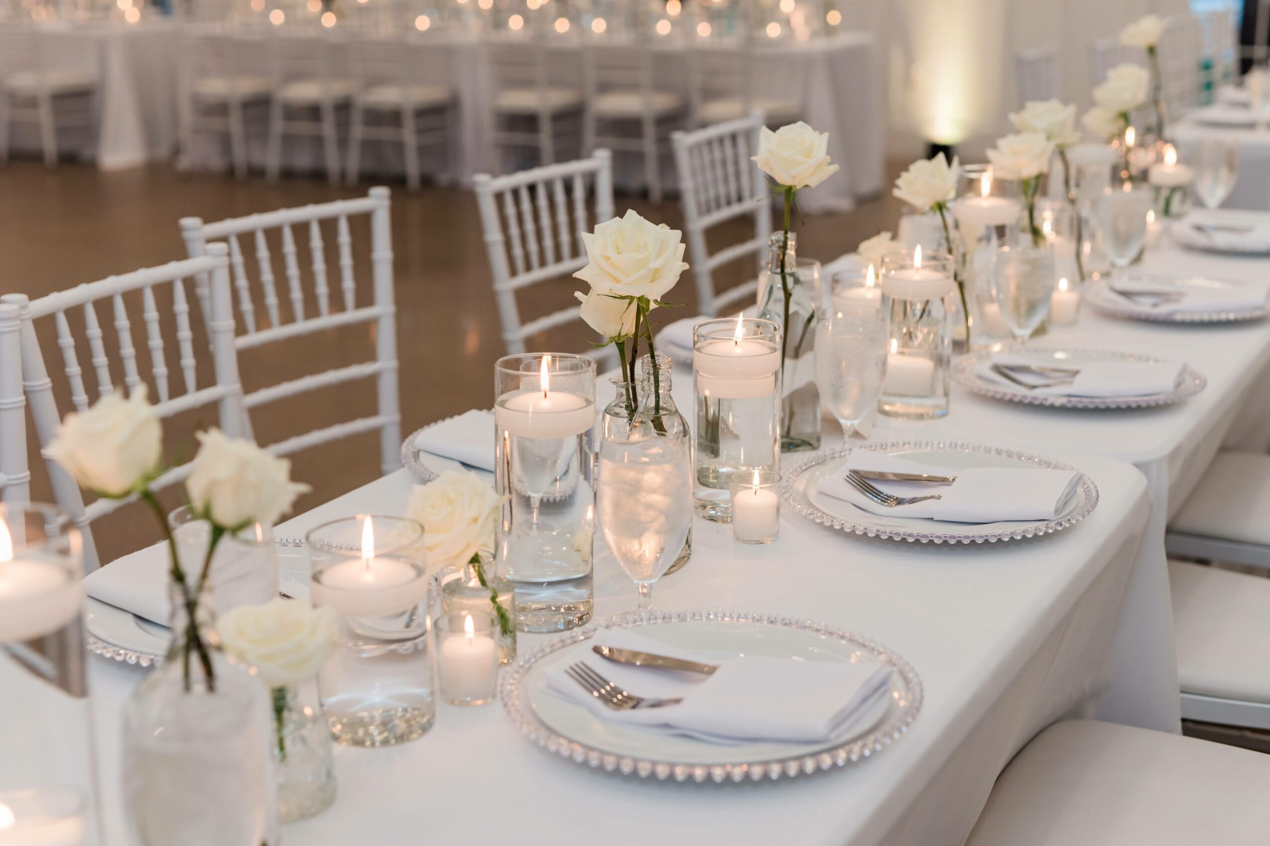 Long banquet table decorated with white roses, candles, and tableware, set for a formal event or wedding reception.