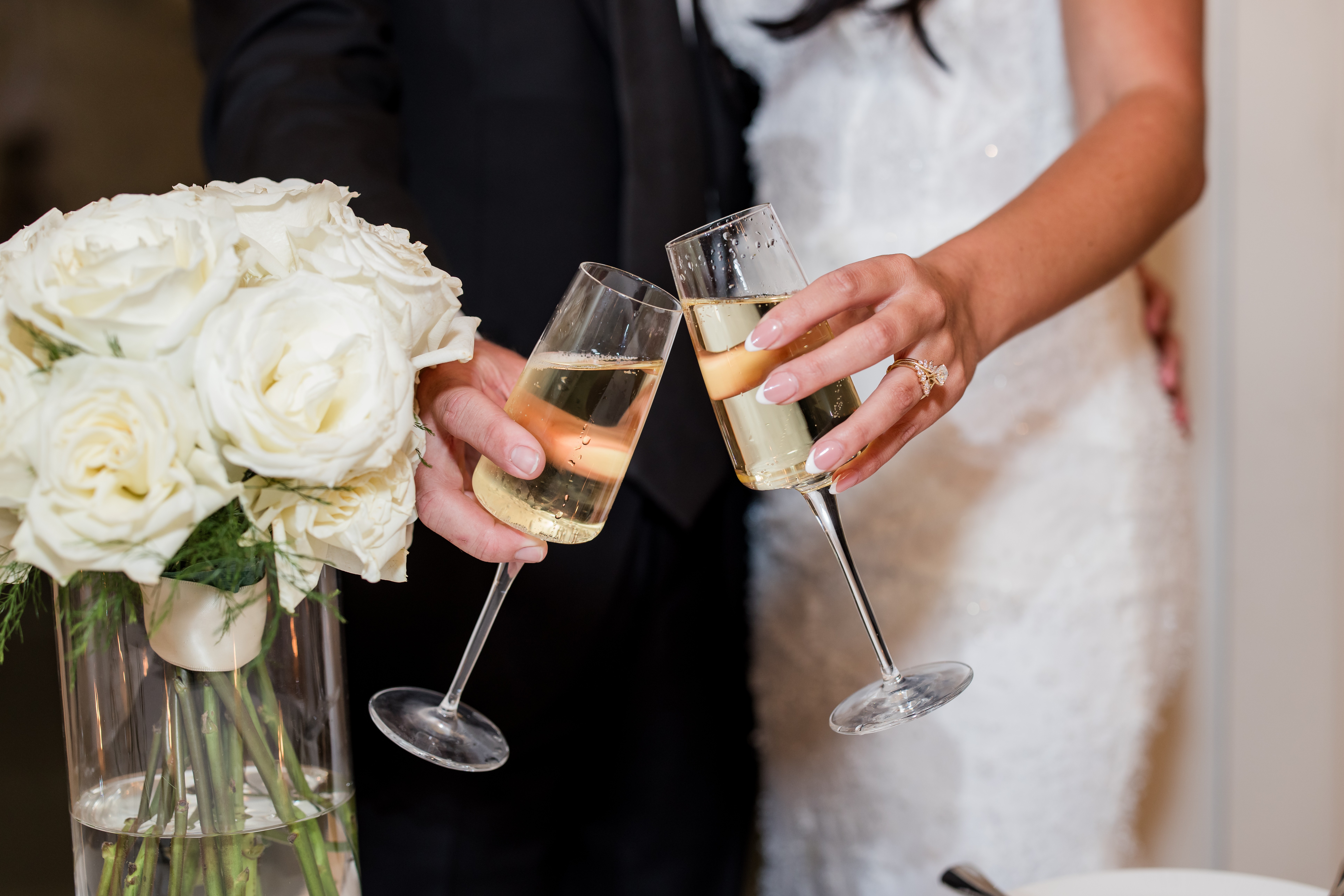 Two people in wedding attire holding champagne glasses, with a bouquet of white roses nearby.