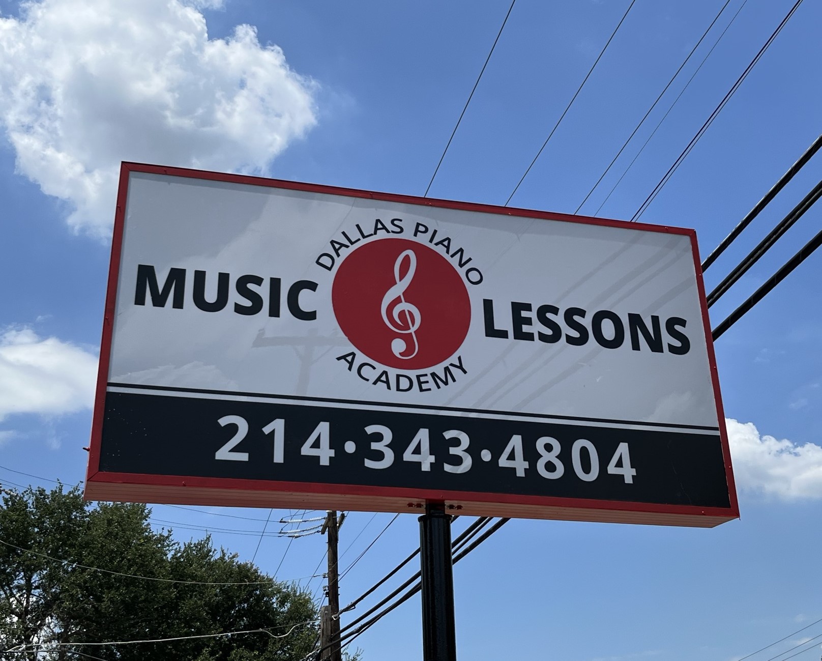 Billboard advertising Dallas Piano Academy music lessons with phone number, against a blue sky with clouds.
