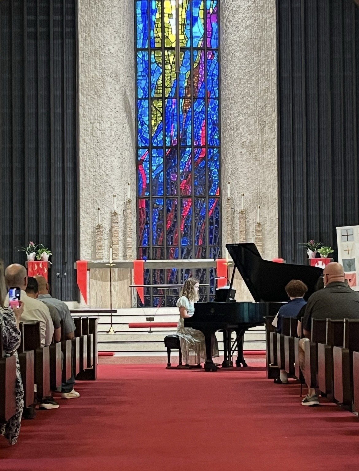 People seated in church pews facing a pianist at a grand piano, with a colorful stained glass window behind. Word count: 20