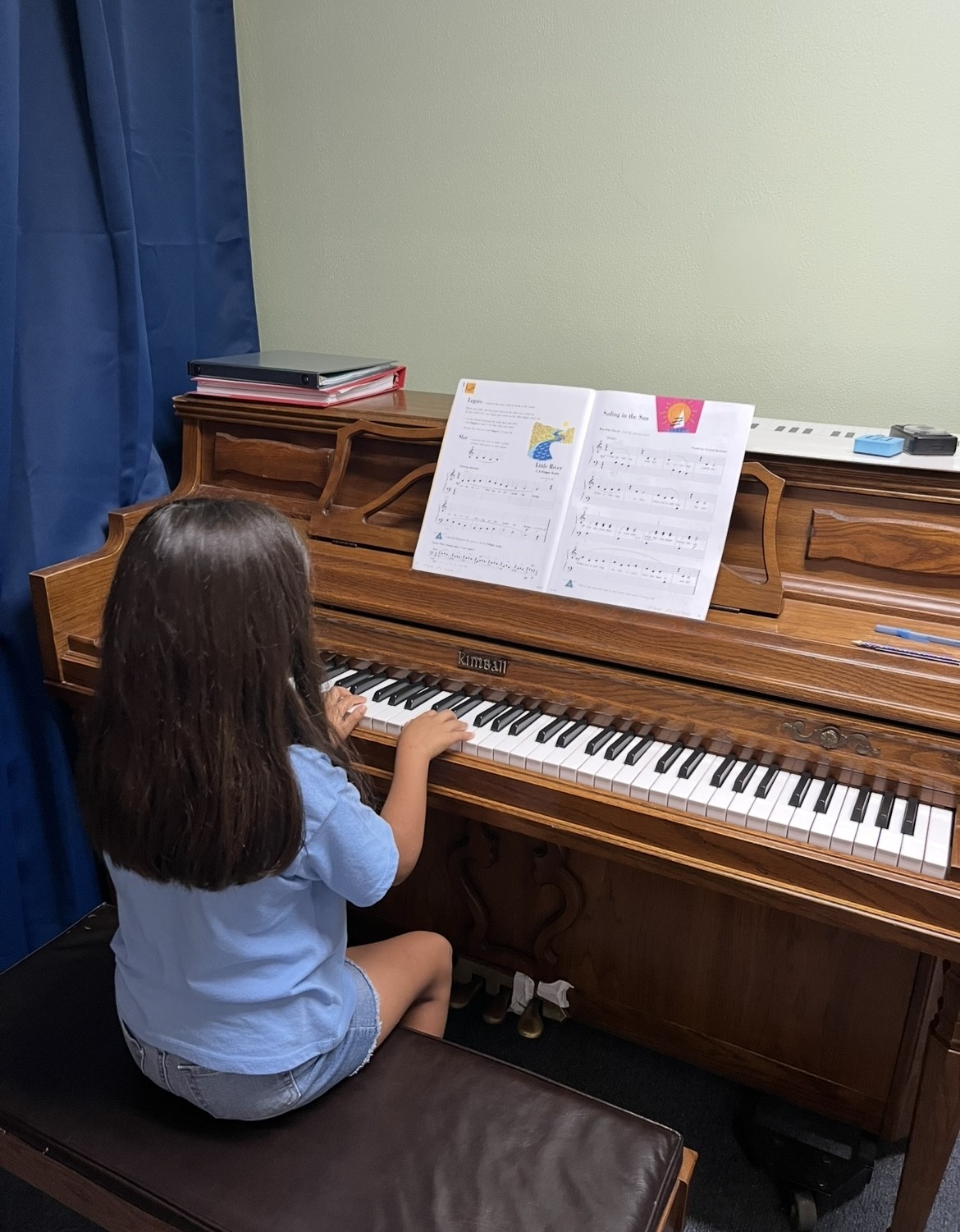 Child with long dark hair sitting at a piano, reading sheet music, in a room with a green wall and blue curtain.
