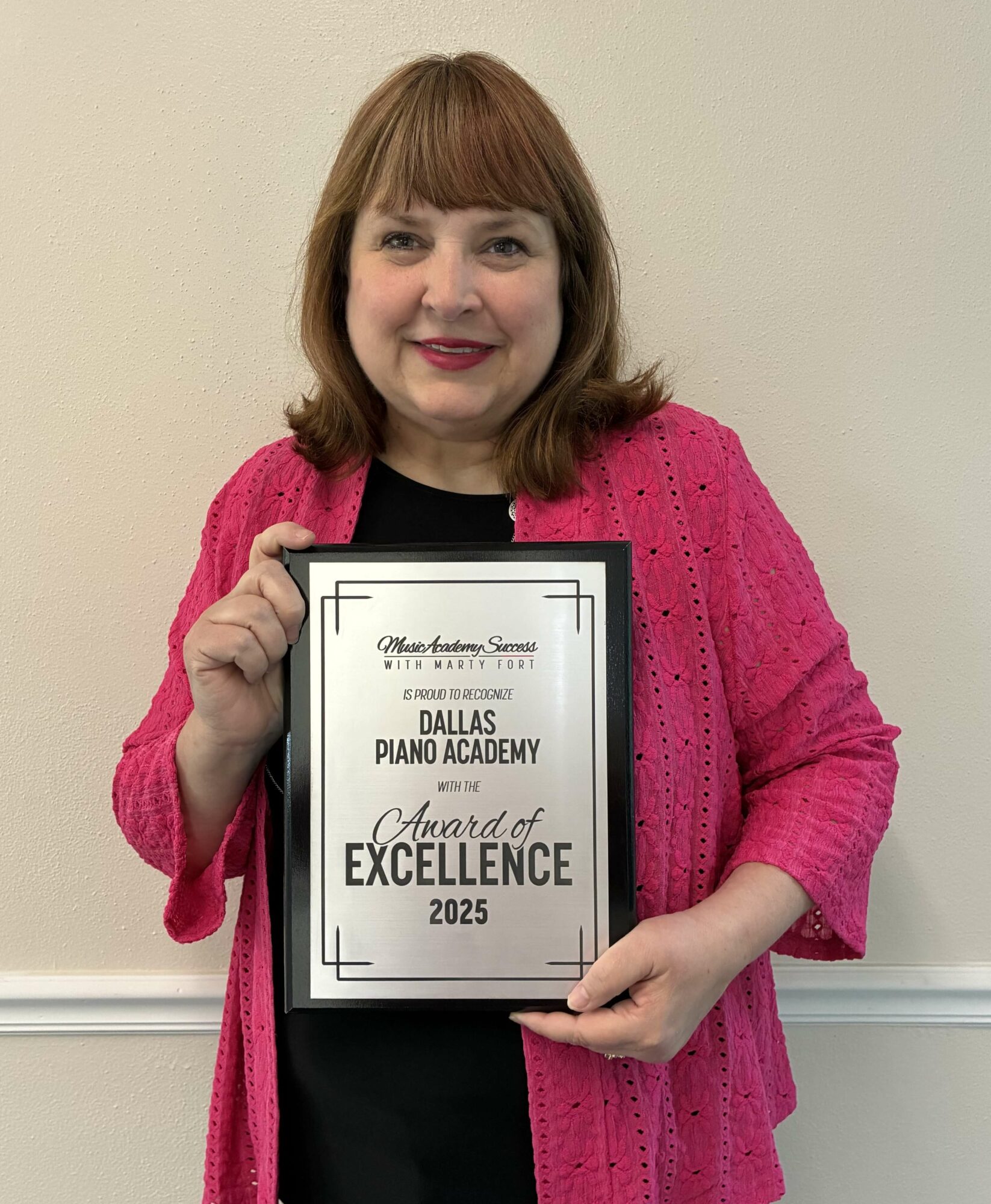 Woman holding award certificate, smiling, wearing pink cardigan and black top, standing against a plain wall.