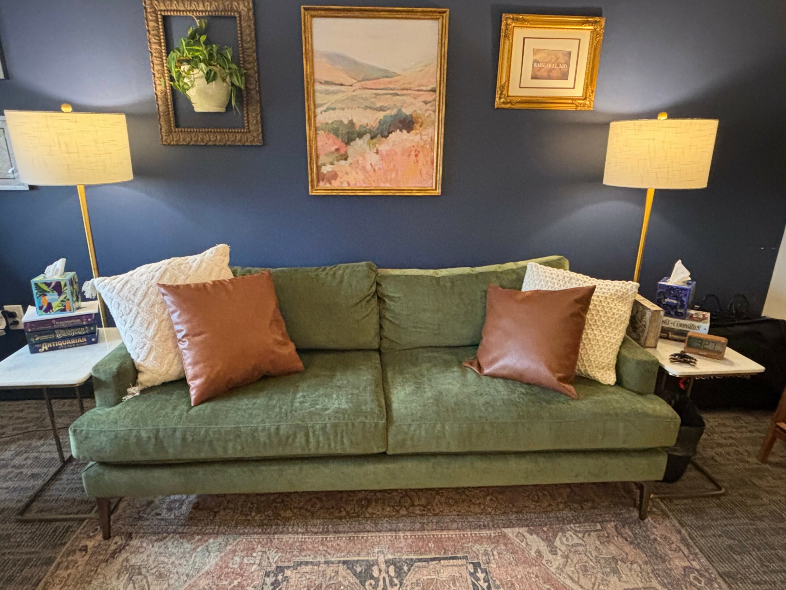 Green sofa with four pillows, two white and two brown, in a living room with wall art and lamps on side tables