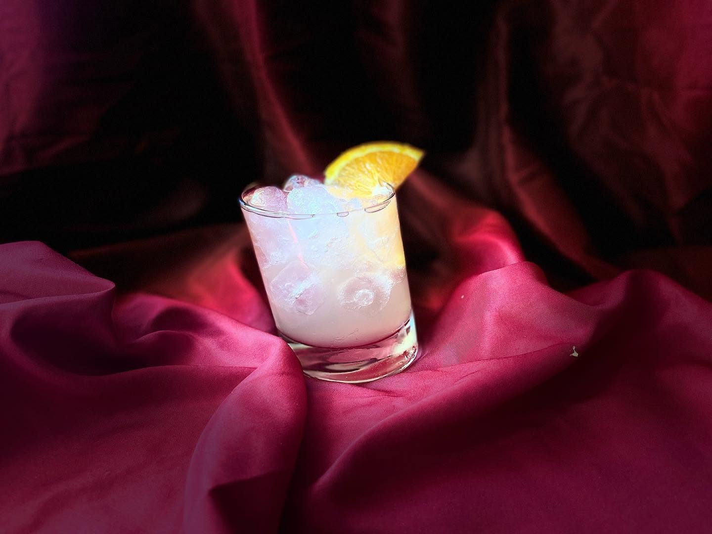 A glass with a lemon wedge on top, filled with a cold beverage and ice, placed on a red fabric background.