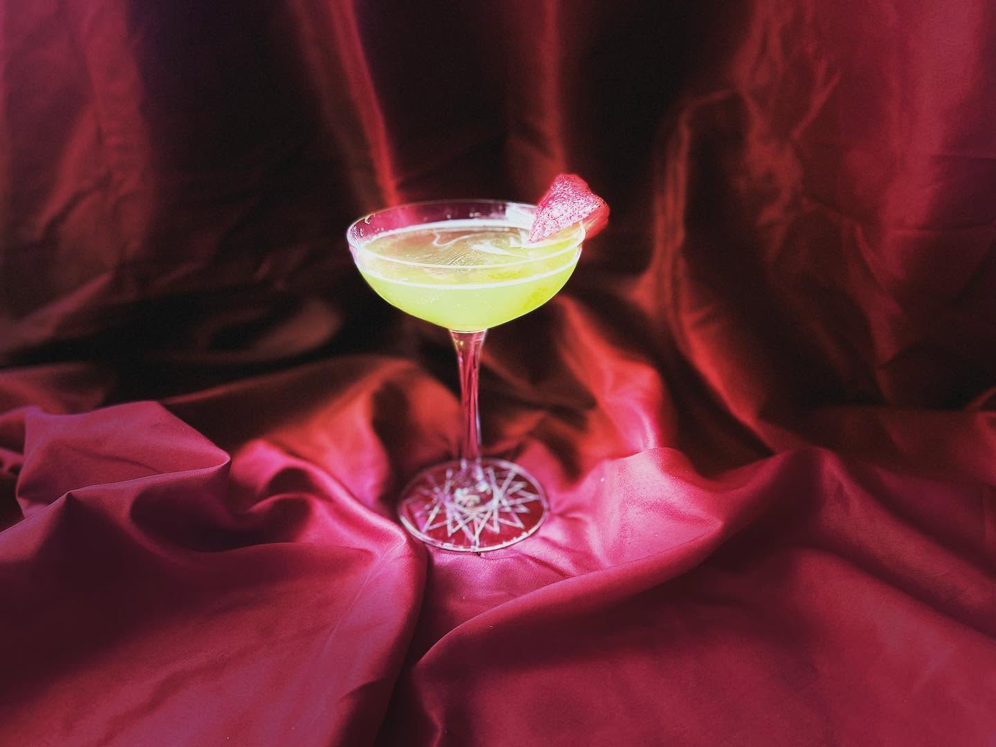 A cocktail glass with a yellow-green drink and a pink garnish, on a red satin fabric background.