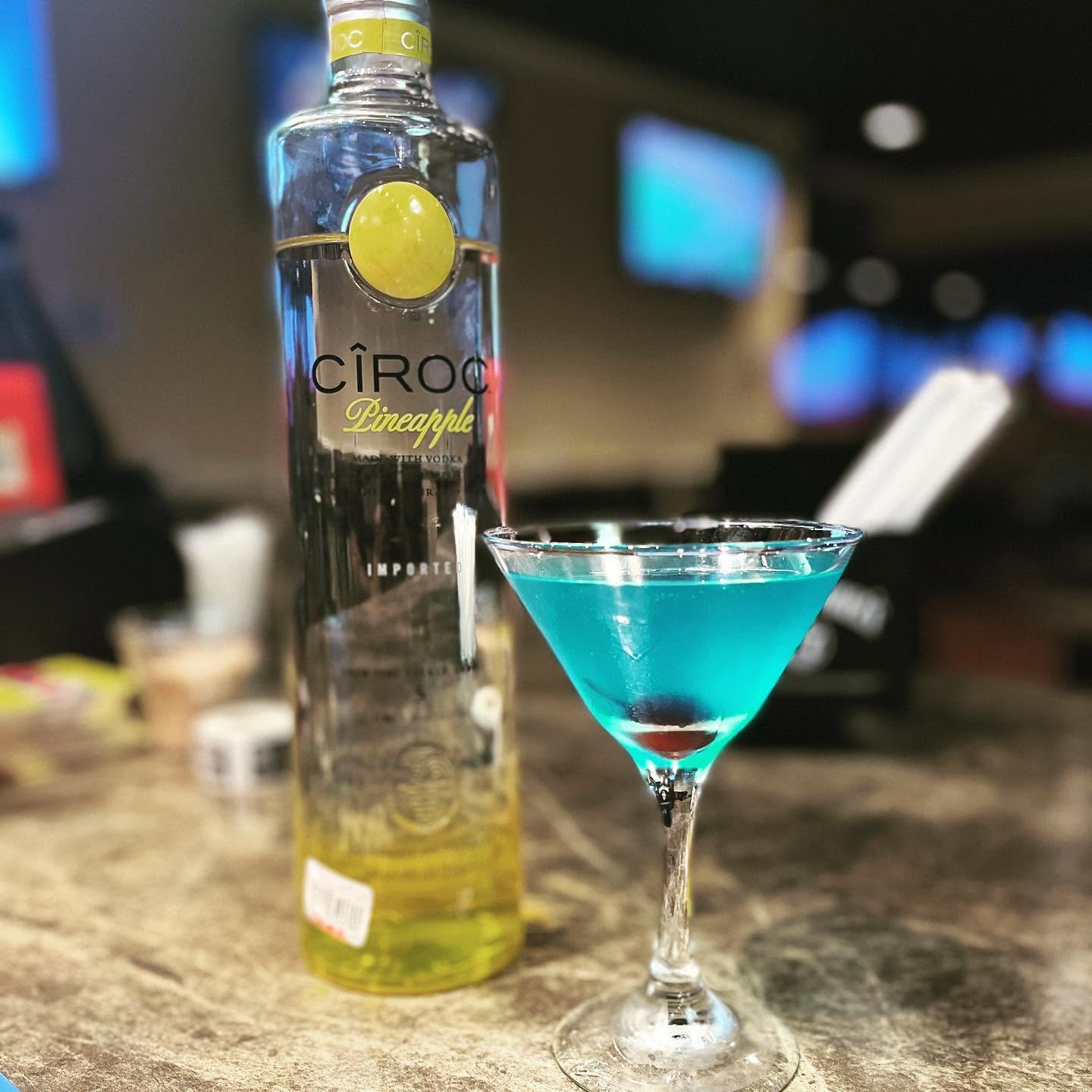 Bottle of Ciroc Pineapple vodka and a blue cocktail in a martini glass on a bar counter.