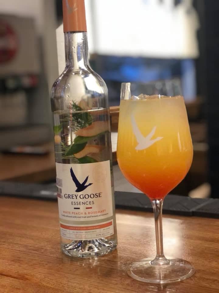 Bottle of Grey Goose Essences and a colorful cocktail with a bird logo on a wooden surface.