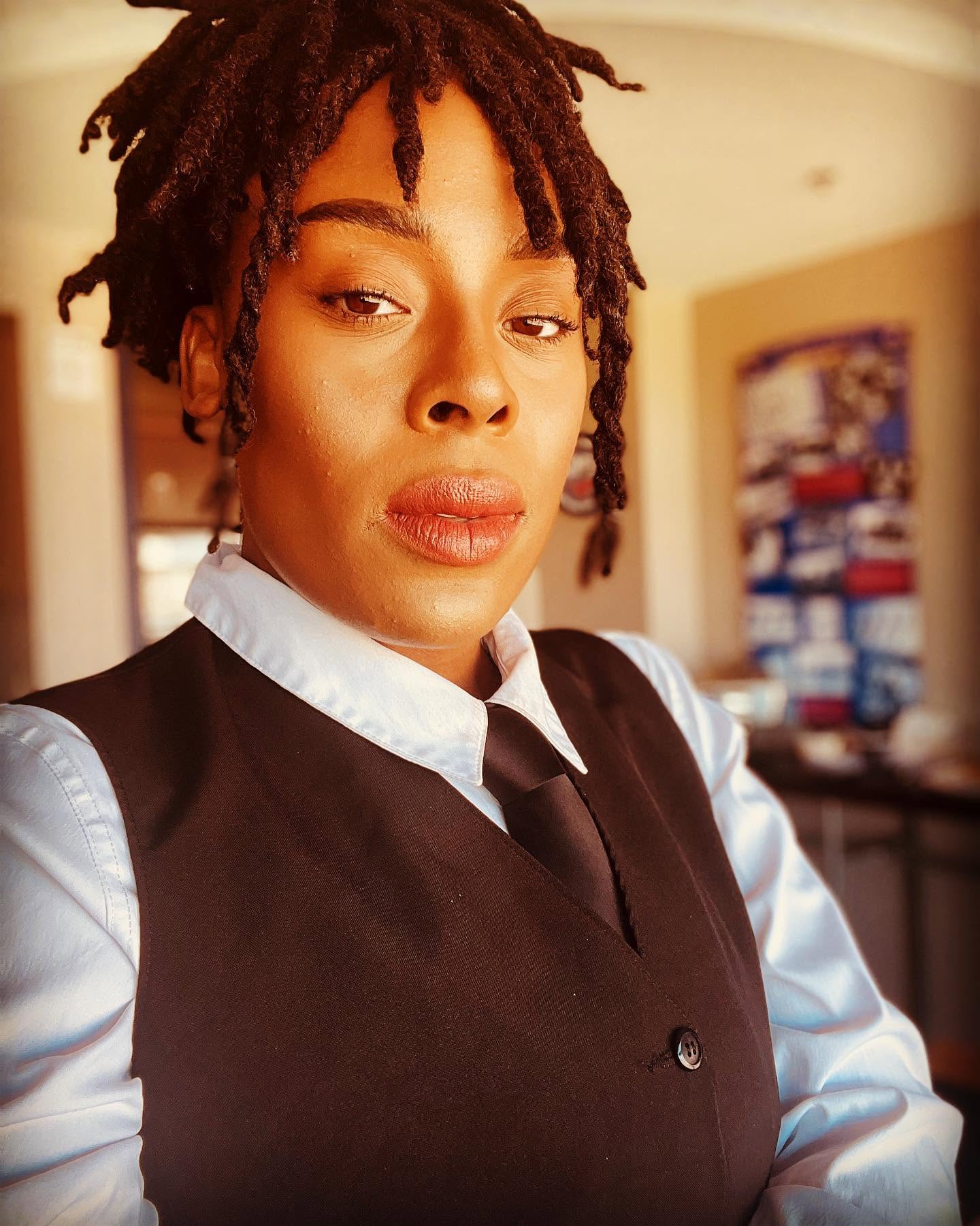 Woman with short dreadlocks wearing a white shirt and black vest, indoors with a blurred background.
