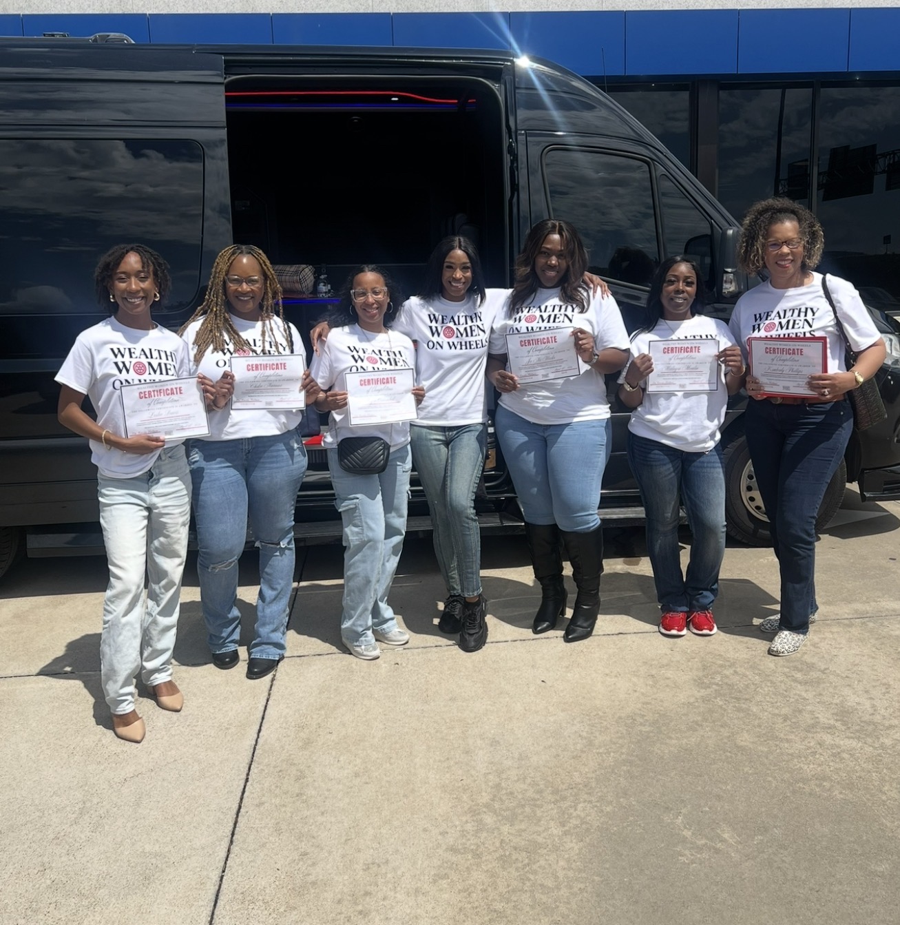 Group of seven women standing in front of a black van, holding signs and certificates, smiling outdoors.