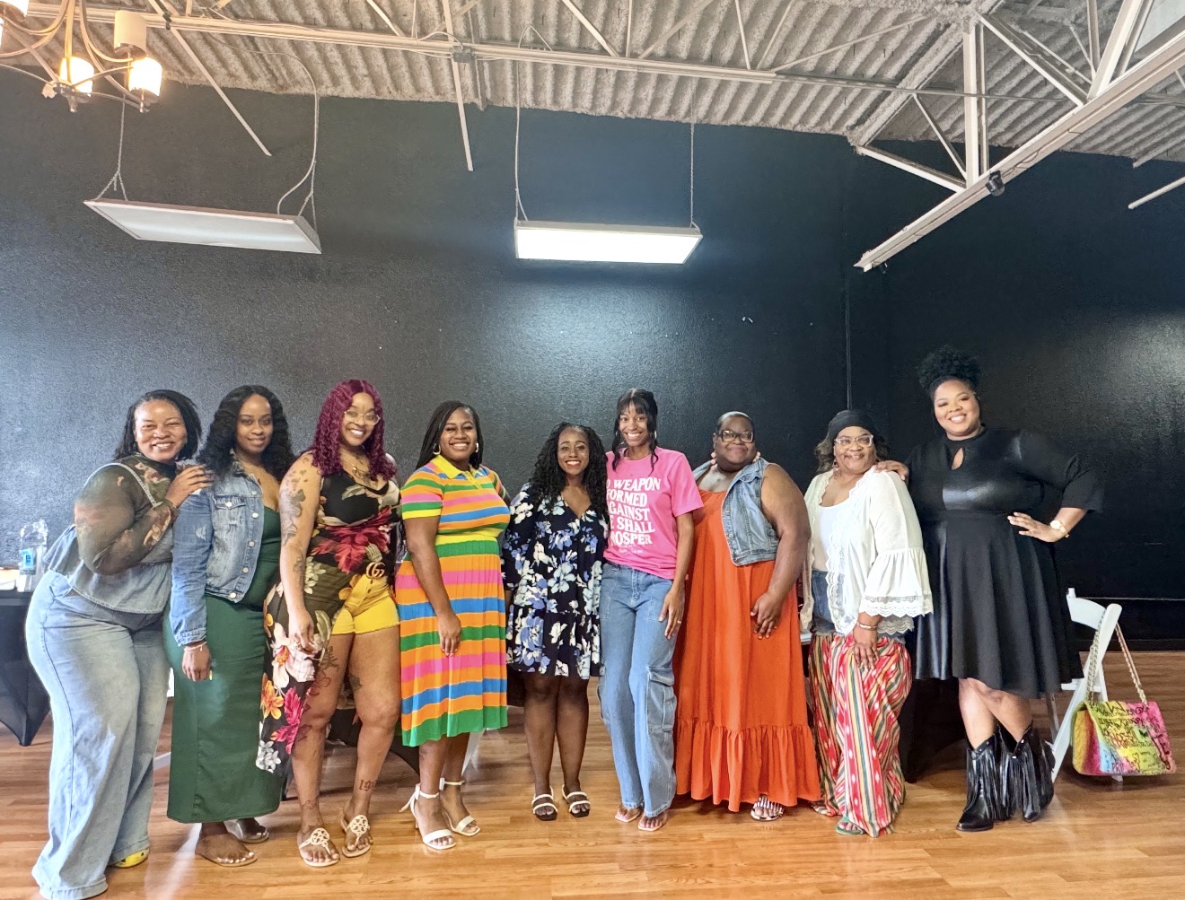 Group of ten women standing in a room with black walls and wooden floor, smiling and posing for a photo.