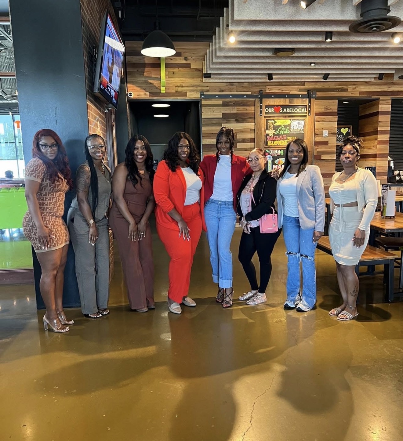 Group of eight women standing indoors, some wearing glasses, in a restaurant or cafe setting with wooden decor.
