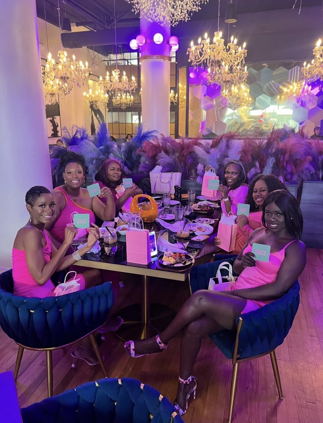 Group of women in pink dresses sitting around a table with drinks and phones at a decorated event venue.