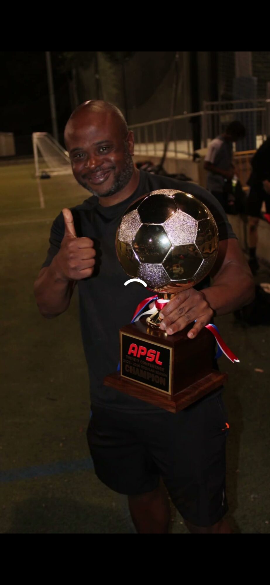 Person smiling and holding a trophy with a soccer ball design, giving a thumbs-up, on a sports field at night.