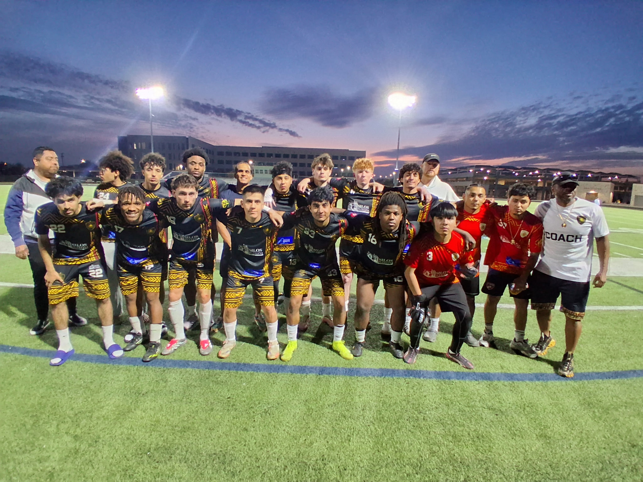 Group of young soccer players and coaches on field at dusk, some in black and yellow uniforms, others in red and black, with stadium lights overhead.