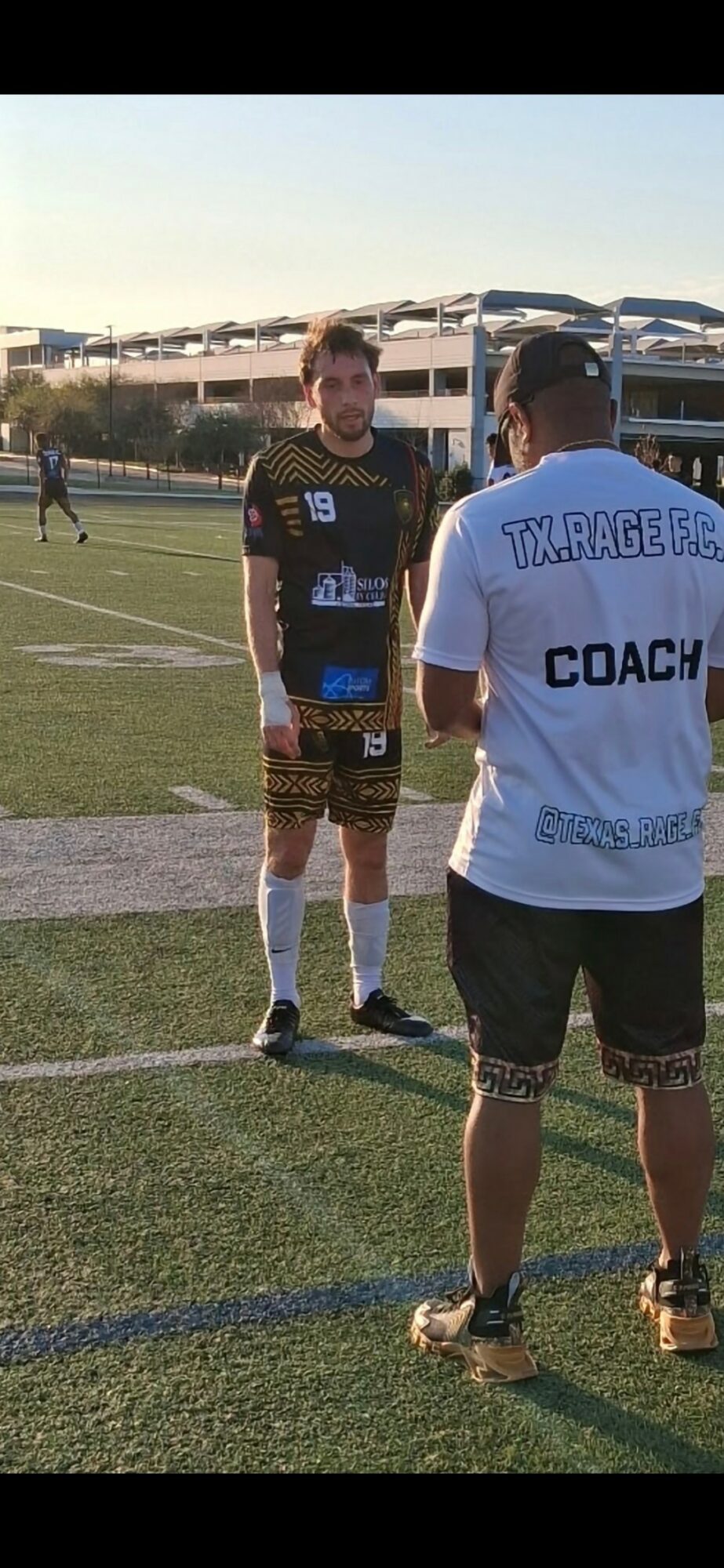 Two men on a soccer field, one wearing a coach shirt, the other in a sports uniform, with a stadium in background.