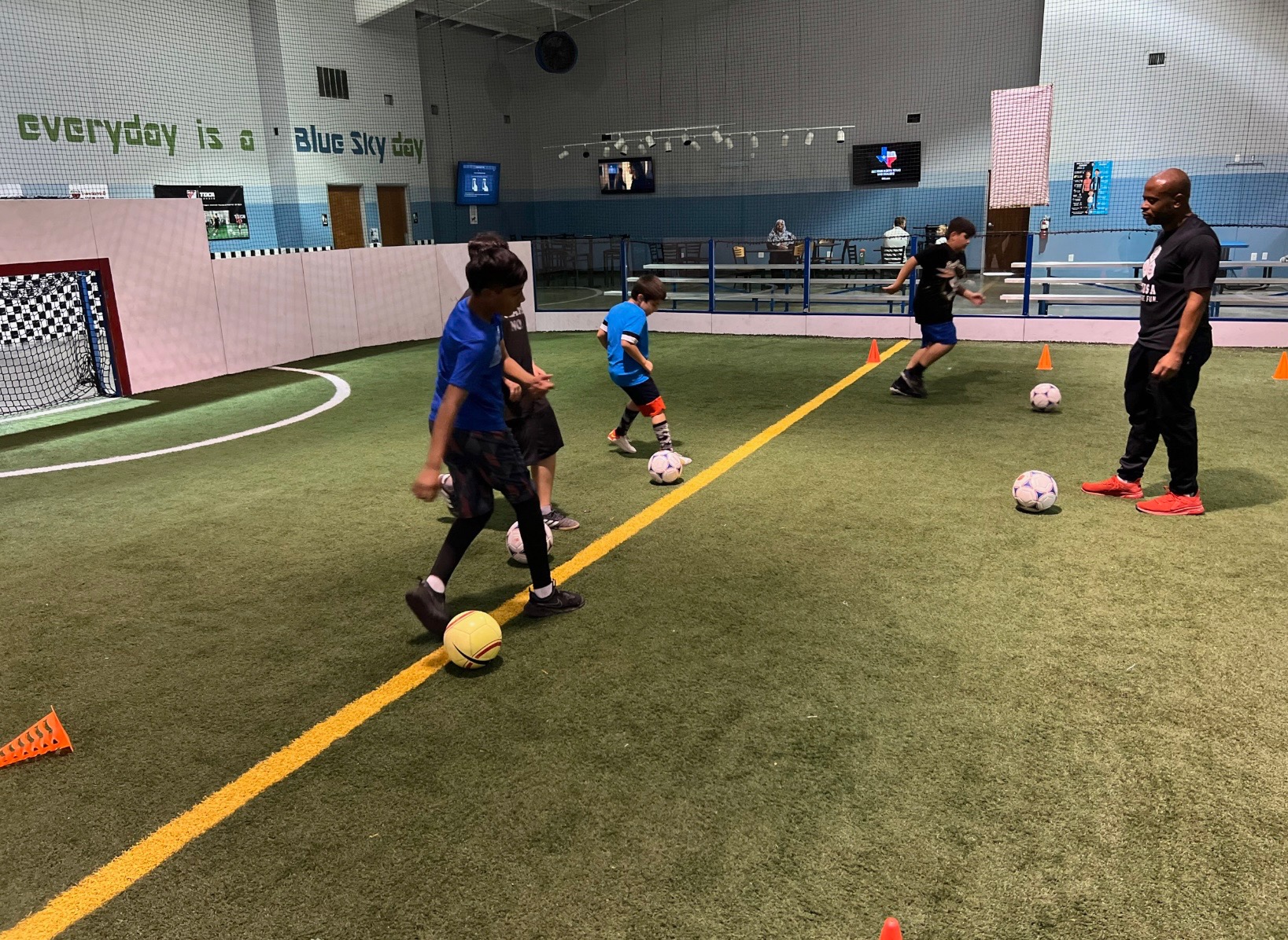 Children and an instructor on an indoor soccer field with soccer balls, practicing dribbling drills.