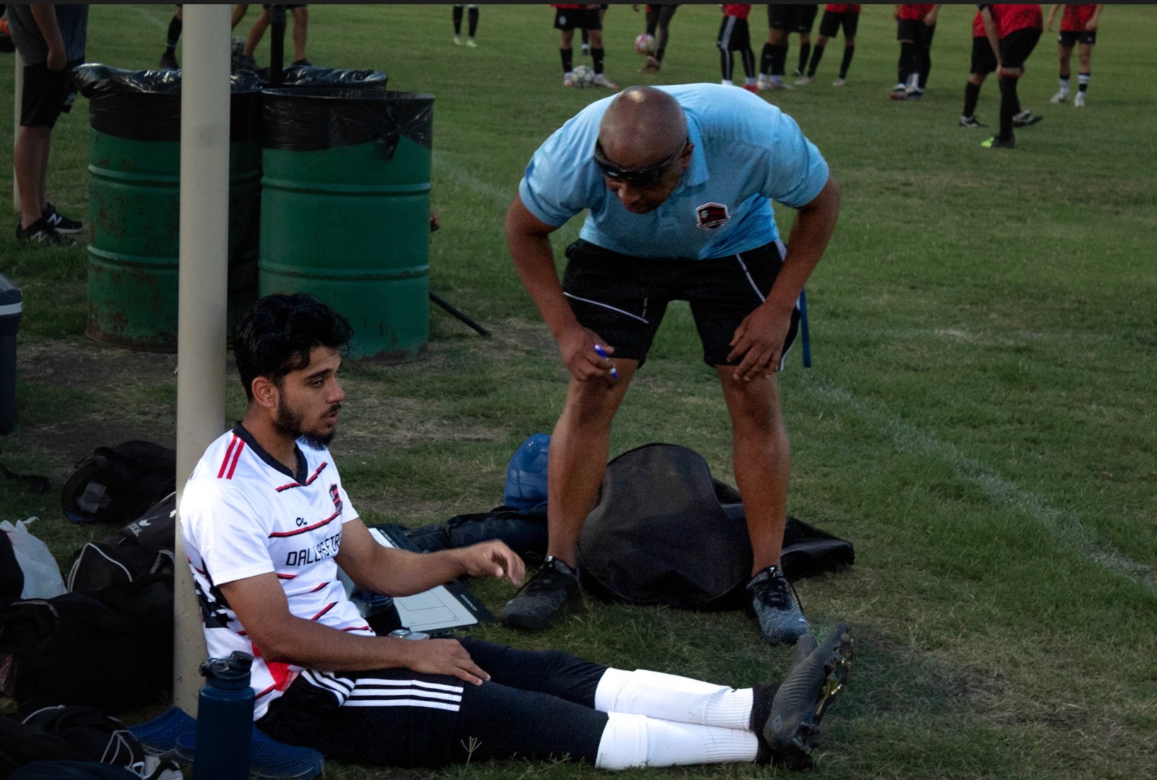 A soccer player sitting on the grass with a man standing nearby, both near sports bags and equipment.