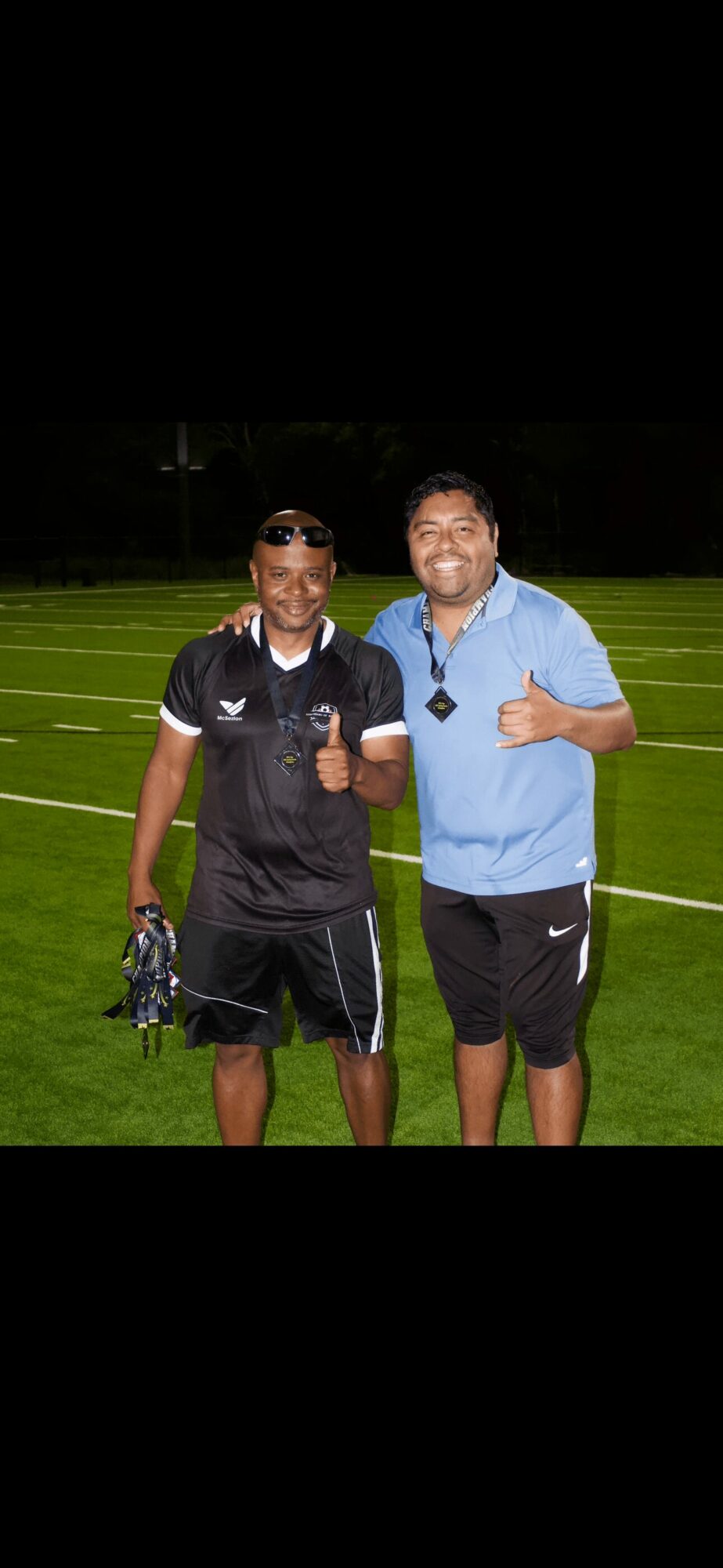 Two men standing on a football field at night, smiling and giving thumbs-up, one in black sportswear and the other in blue, holding a whistle.