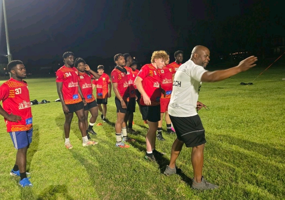 Group of soccer players in red jerseys standing on a grassy field, listening to a coach in white shirt and black shorts.