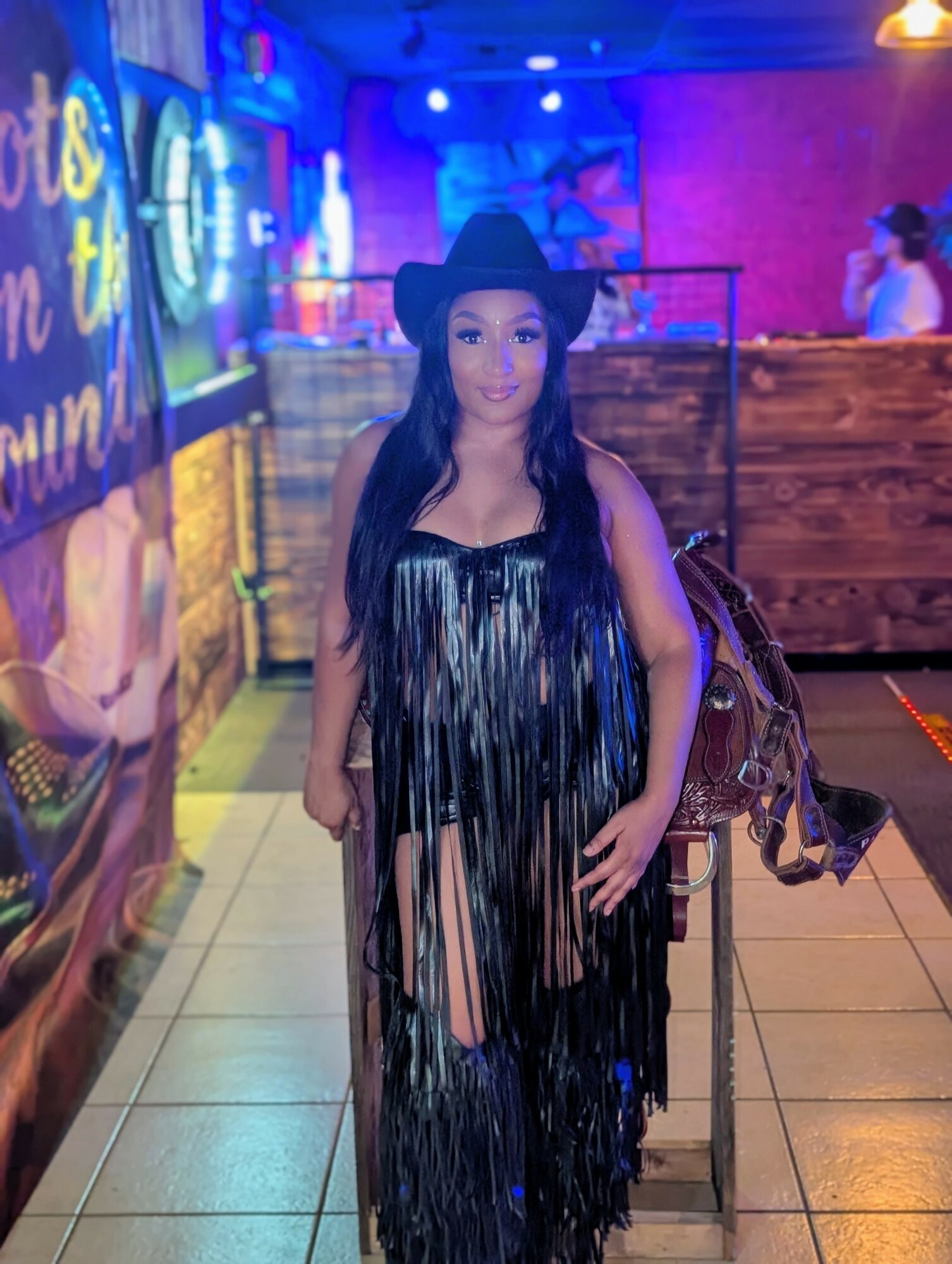 Woman in a black hat and fringed dress standing in a colorful indoor setting with neon lights.
