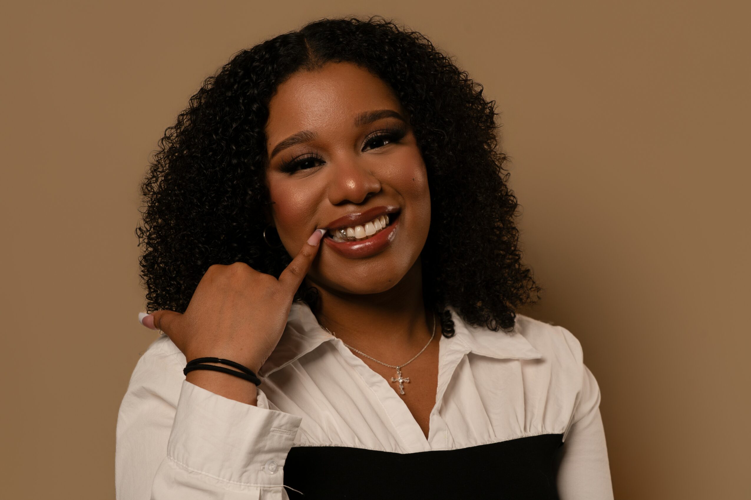 Smiling woman with curly hair wearing a white shirt and black top, touching her face, against a beige background.
