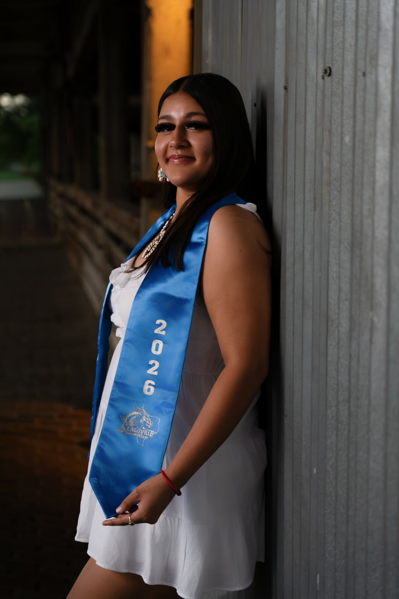 Young woman in white dress with blue sash, smiling, standing against a corrugated metal wall outdoors.
