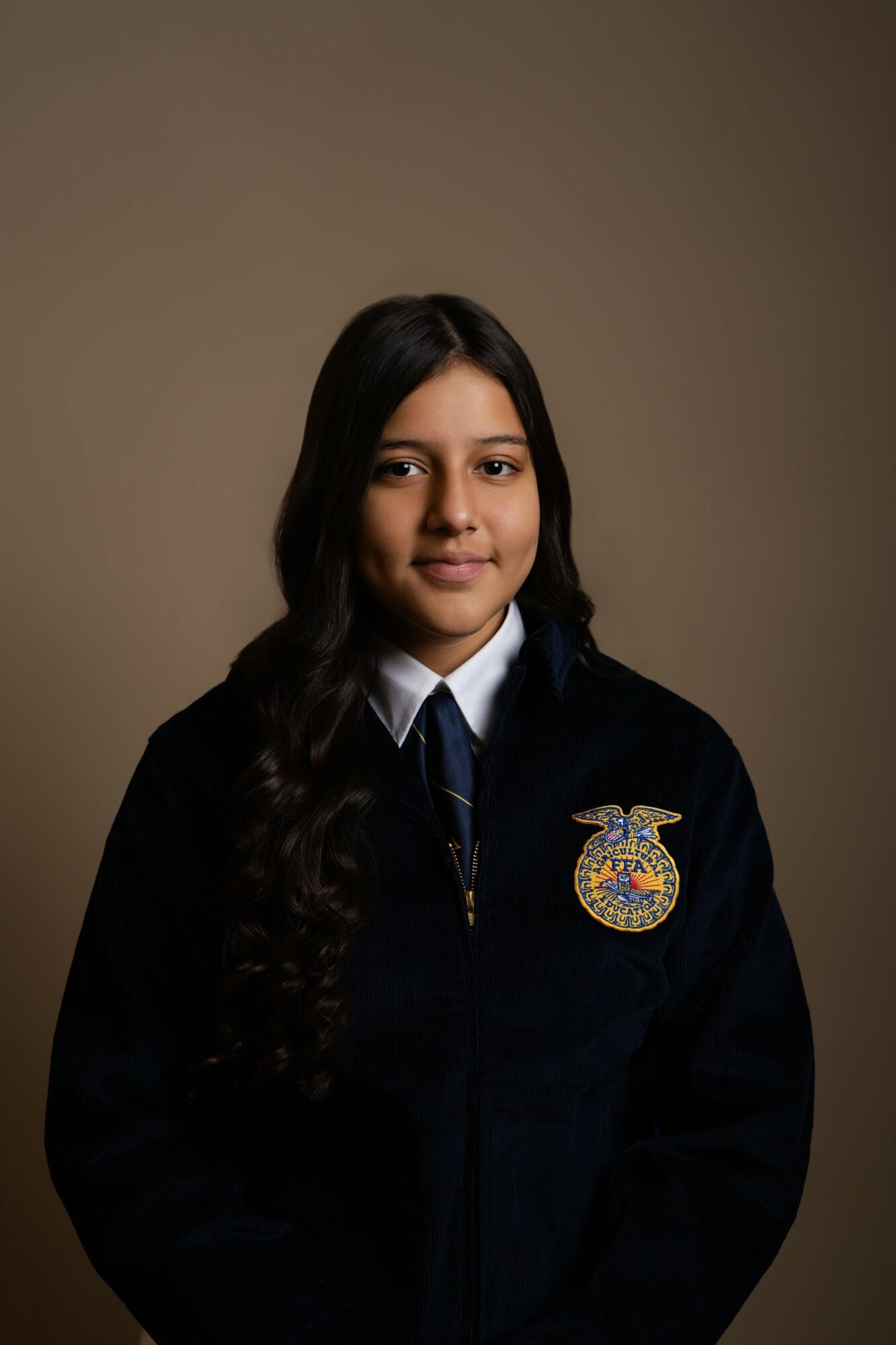 Young woman with long dark hair wearing a dark jacket with an embroidered emblem, white shirt, and tie, against a plain background.
