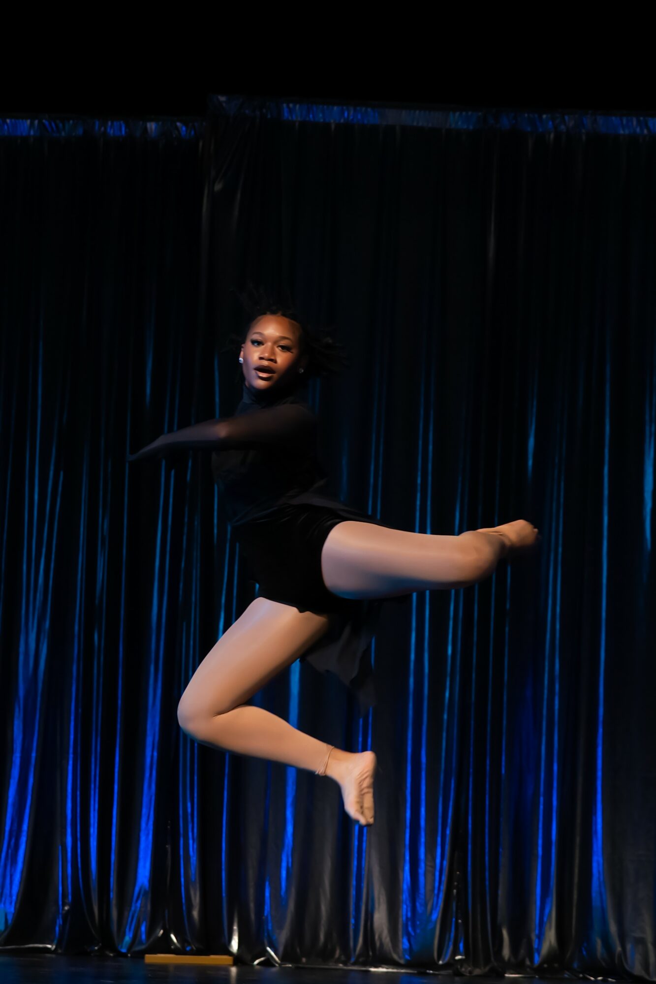 Young girl performing a dance move on stage with dark curtains in background.
