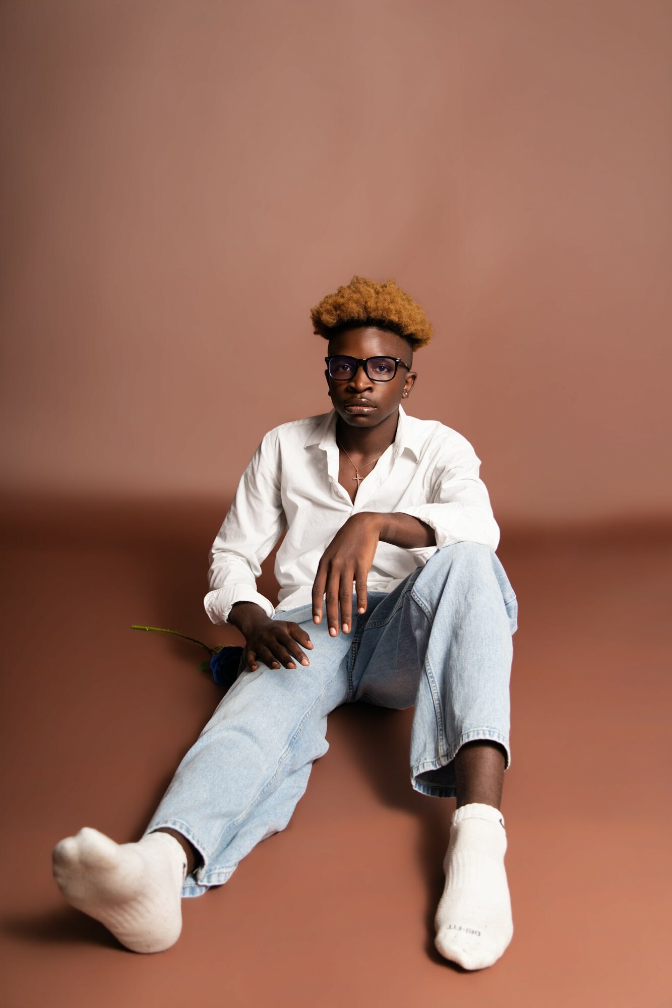 Person with curly blonde hair and glasses sitting on the floor, wearing a white shirt, light blue jeans, and white shoes, against a brown background.