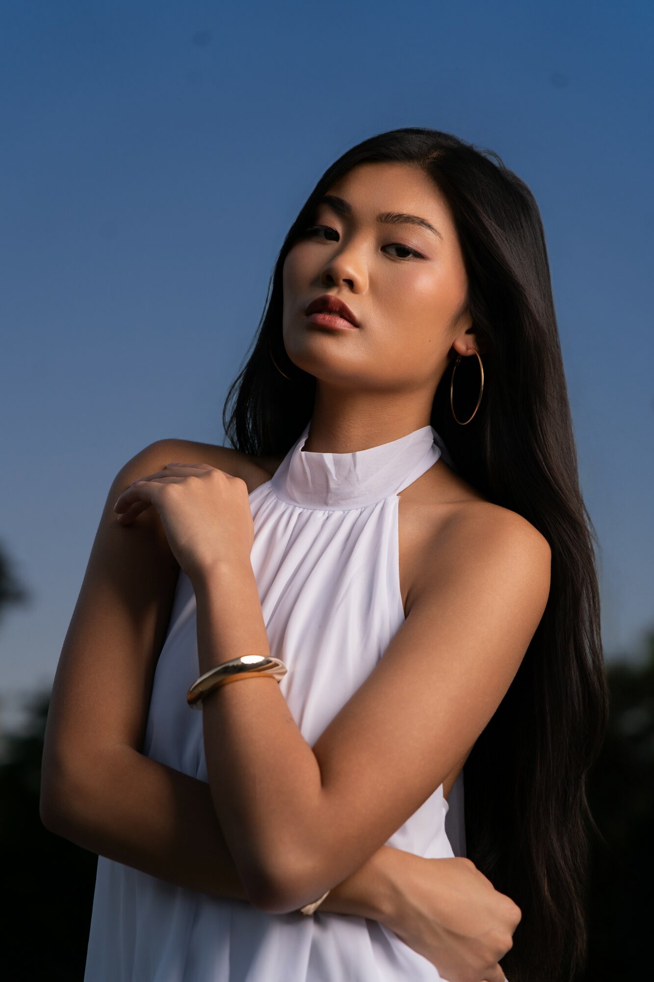 Young woman with long dark hair wearing a white sleeveless top and hoop earrings, posing outdoors against a blue sky.
