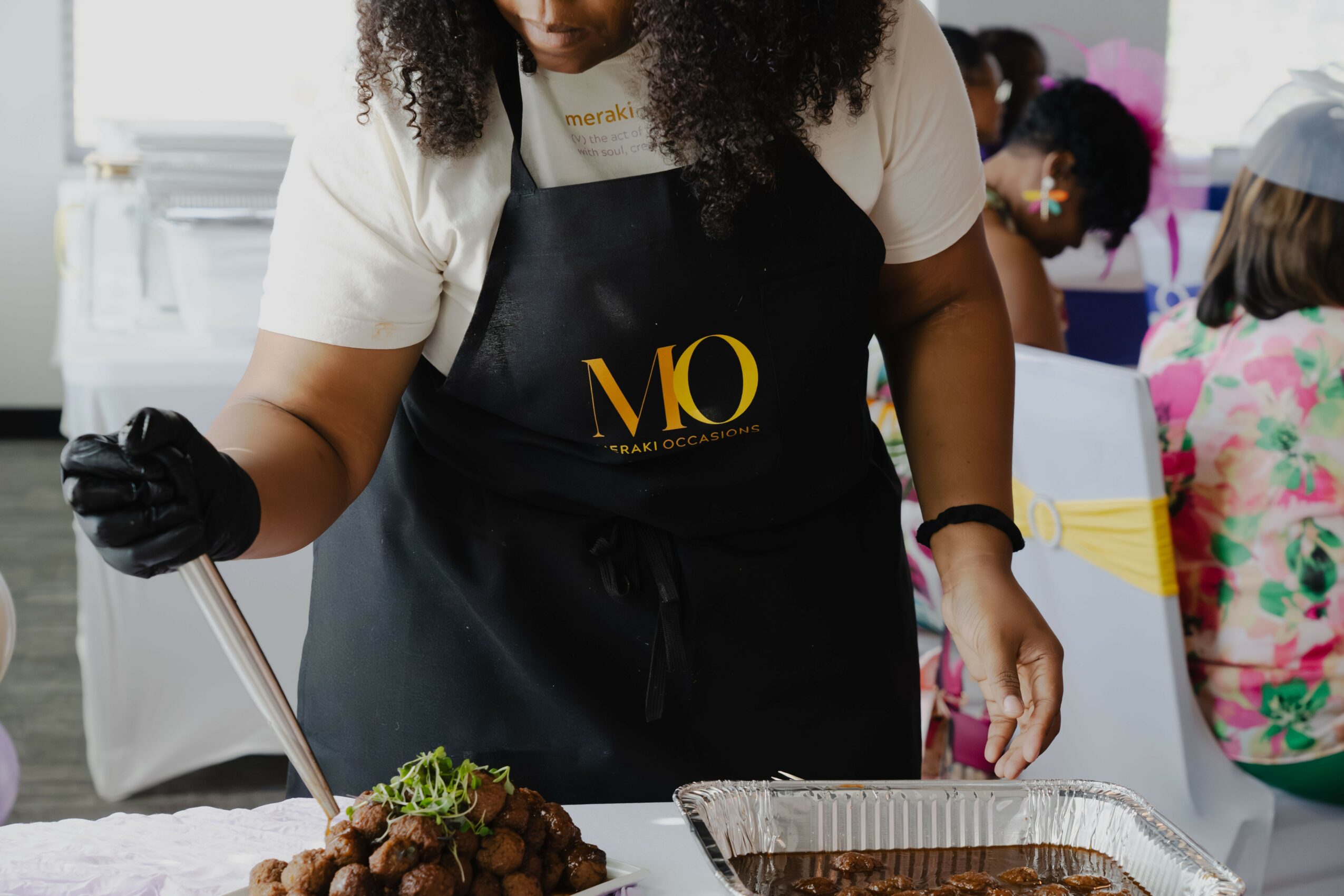 Person wearing a black apron preparing food at a table, with children in colorful clothing in the background.