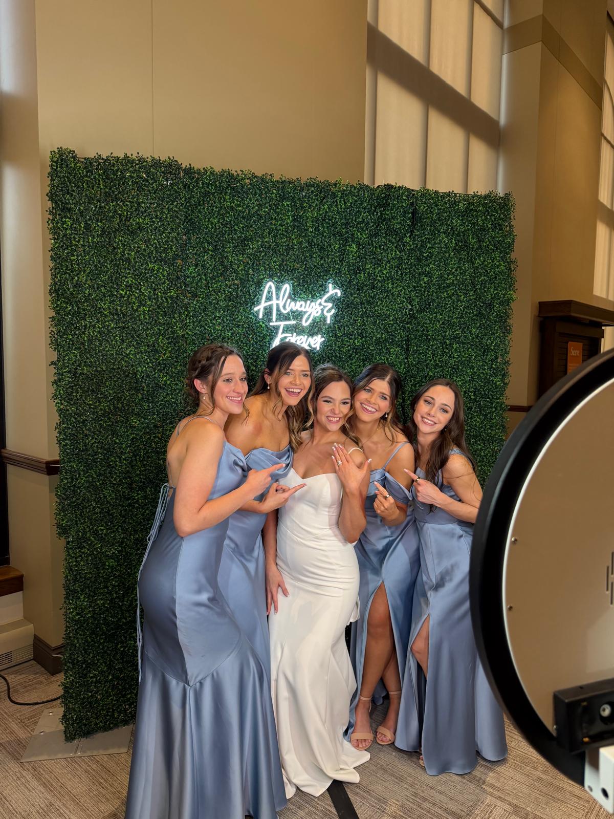 Five women in formal dresses standing in front of a green hedge wall with illuminated sign, smiling, posing for photo.