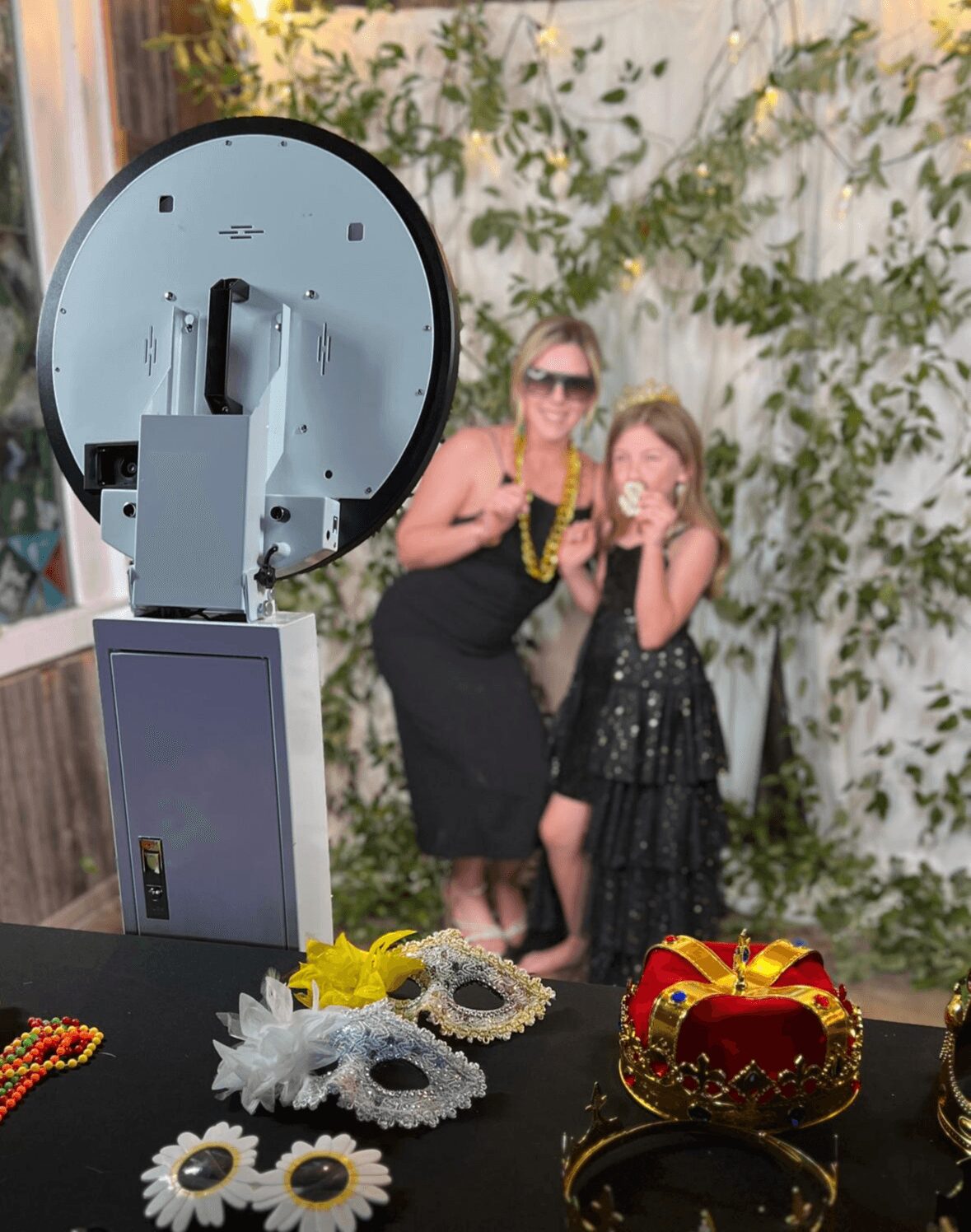 Two women, one adult and one child, smiling and posing outdoors with a floral background, wearing sunglasses and black dresses.