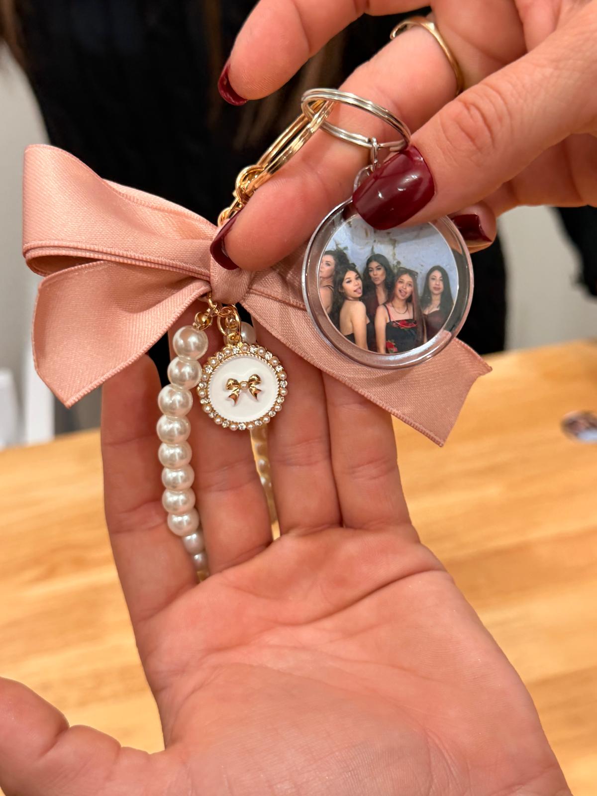 Hand holding keychain with a pink bow, pearl charm, and a round photo of four women, against a wooden surface.