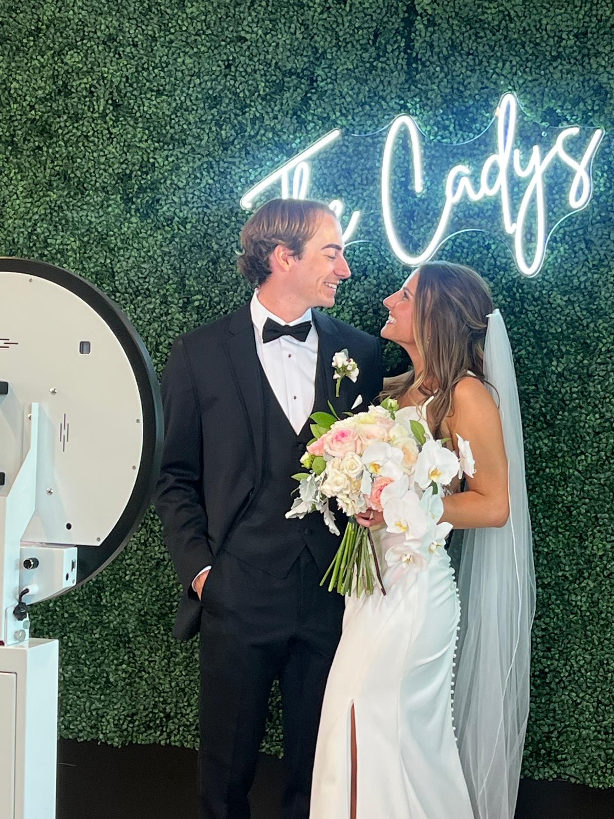 Bride and groom looking at each other, bride holding a bouquet, wedding sign in background, green wall backdrop.