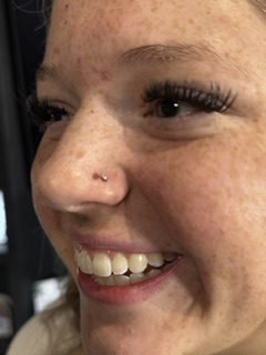 Close-up of a smiling person's face showing freckles, eye makeup, and a nose piercing.
